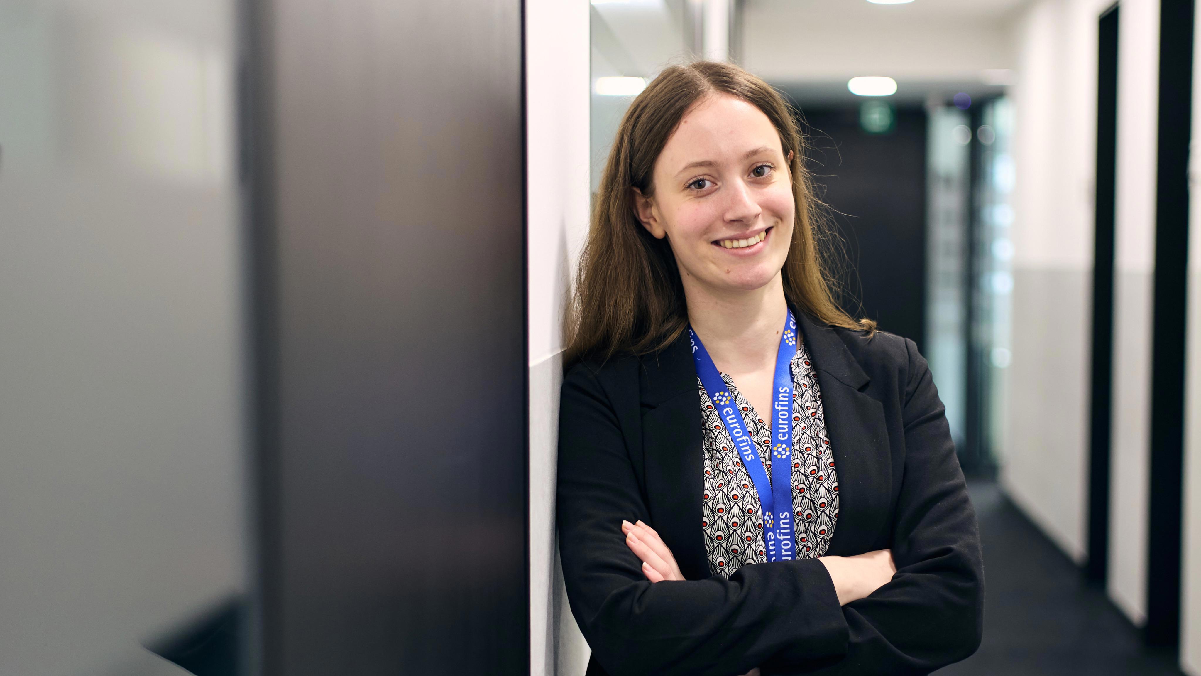 A woman leaning against a wall in an office corridor with her arms crossed.  The woman is smiling directly at the camera and is wearing a lanyard, blouse and blazer. 