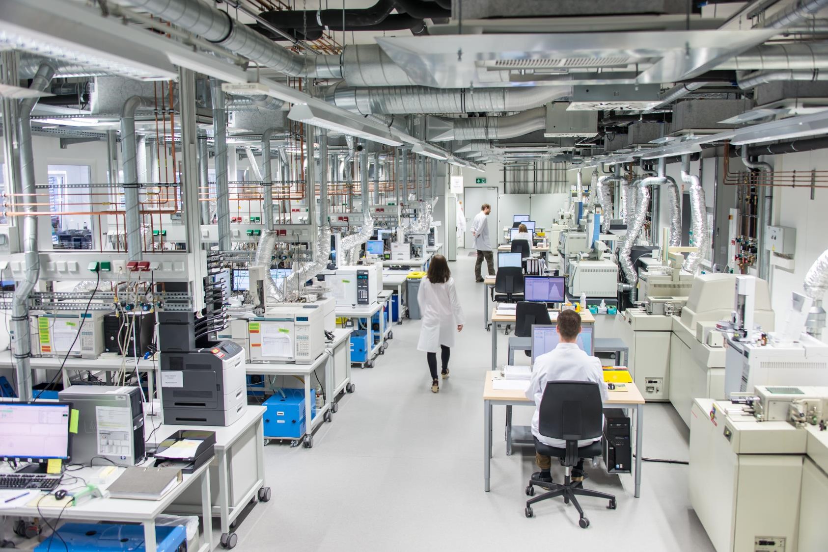 A modern laboratory with advanced equipment and four people in lab coats working. Two people are at computers, while one person is walking through the laboratory and another is standing up looking at a computer. The room is well-lit with numerous machines and scientific instruments arranged neatly.