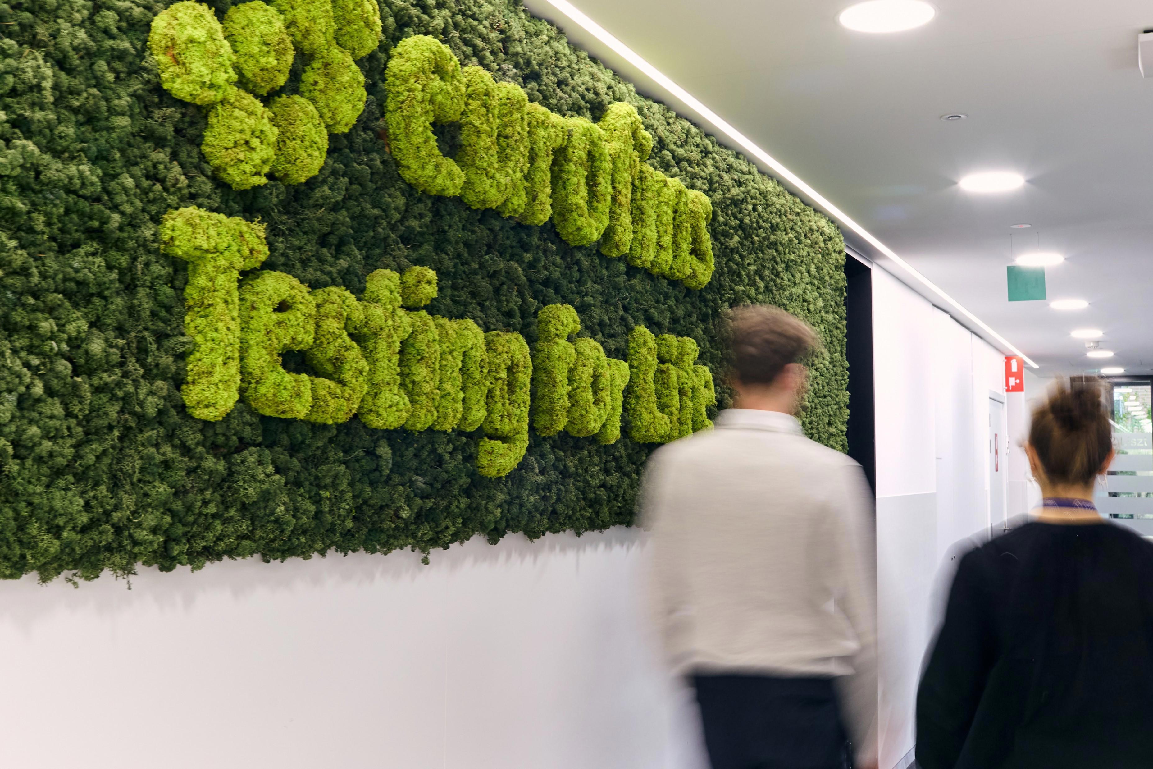 Two people walking down a hallway with a green moss wall on the left side. The moss wall has the words "Eurofins Testing for Life" embedded in it. The hallway is well-lit with white walls and ceiling lights