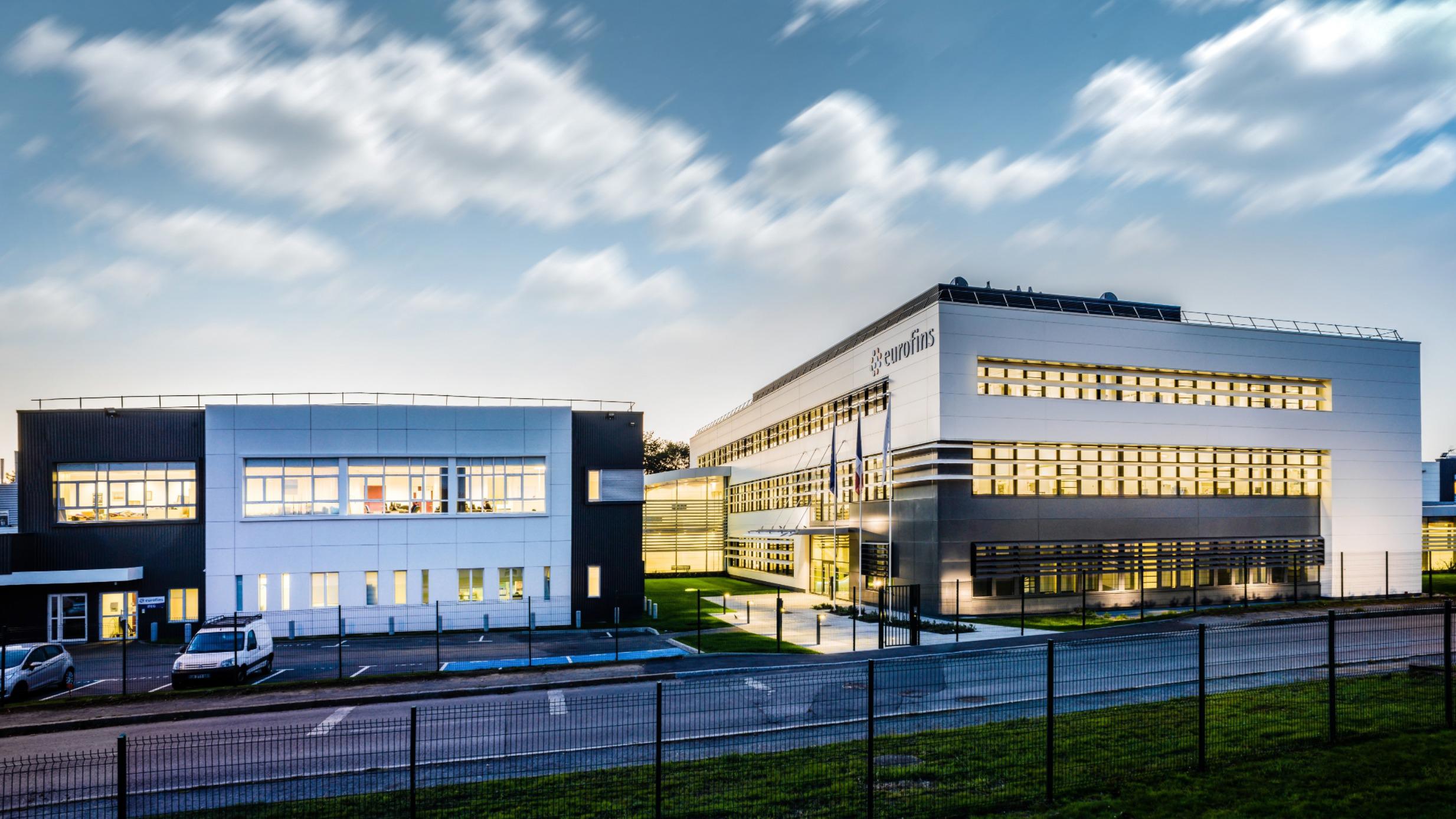 The Eurofins laboratory in Nantes, France, featuring two modern buildings with large windows, illuminated during twilight. The buildings are surrounded by a well-maintained lawn and a fence in the foreground. The sky is partly cloudy, and several cars are parked near the buildings.