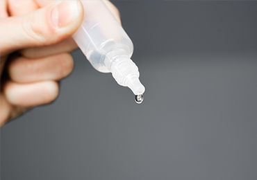 Close-up image of a hand holding a small plastic bottle with a dropper tip, used for oral and ear liquid formulations. A single drop of liquid is about to fall from the tip, with a plain, blurred background emphasizing the droplet.