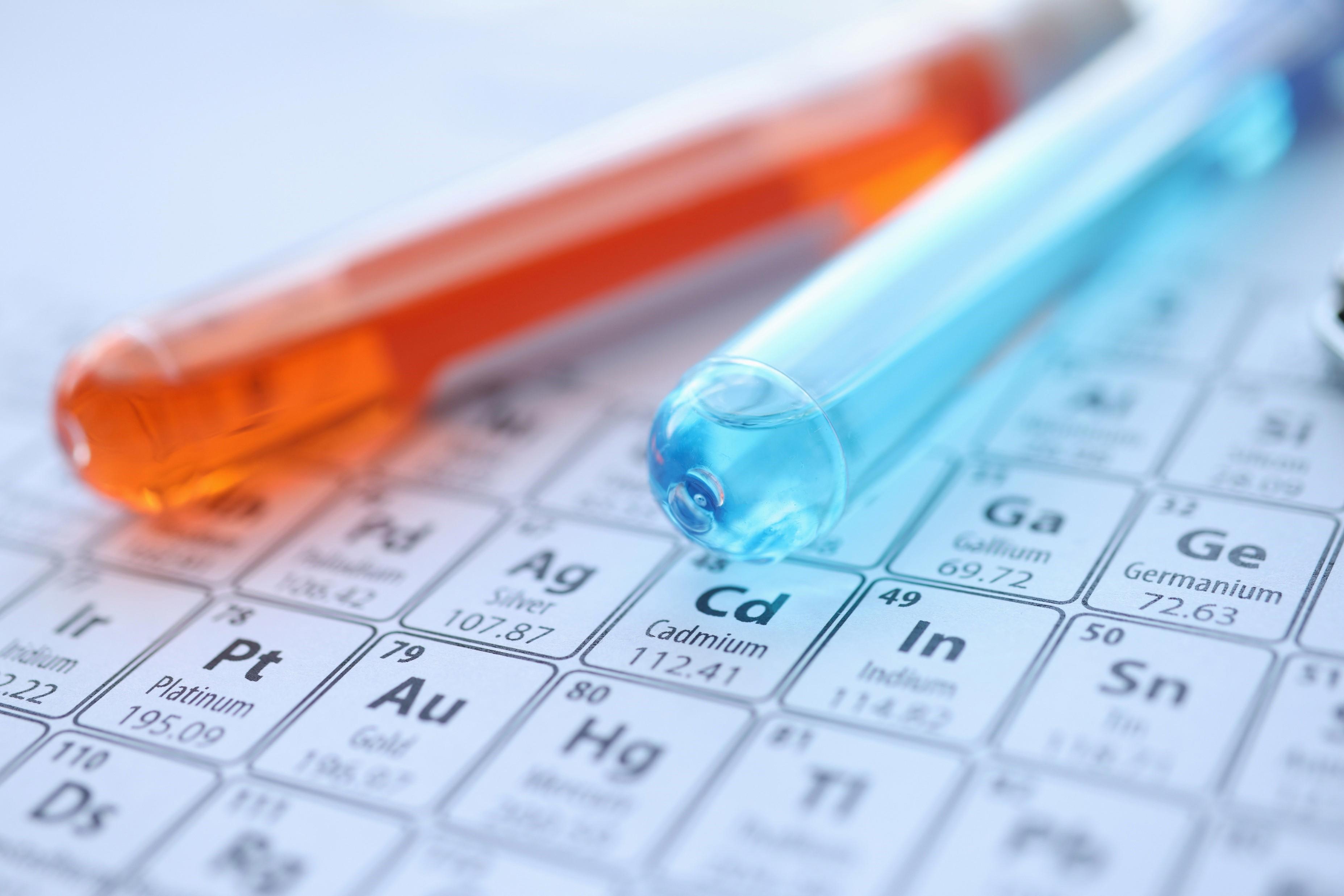 Two test tubes, one containing an orange liquid and the other containing a blue liquid, are placed on top of a periodic table of elements. The periodic table is partially visible, showing elements such as Cadmium (Cd), Silver (Ag), Platinum (Pt), Gold (Au), Mercury (Hg), and Indium (In).
