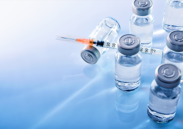 Several clear glass vials with metal caps, filled with a transparent liquid, arranged on a reflective blue surface. One vial is lying on its side with a syringe partially inserted into it.