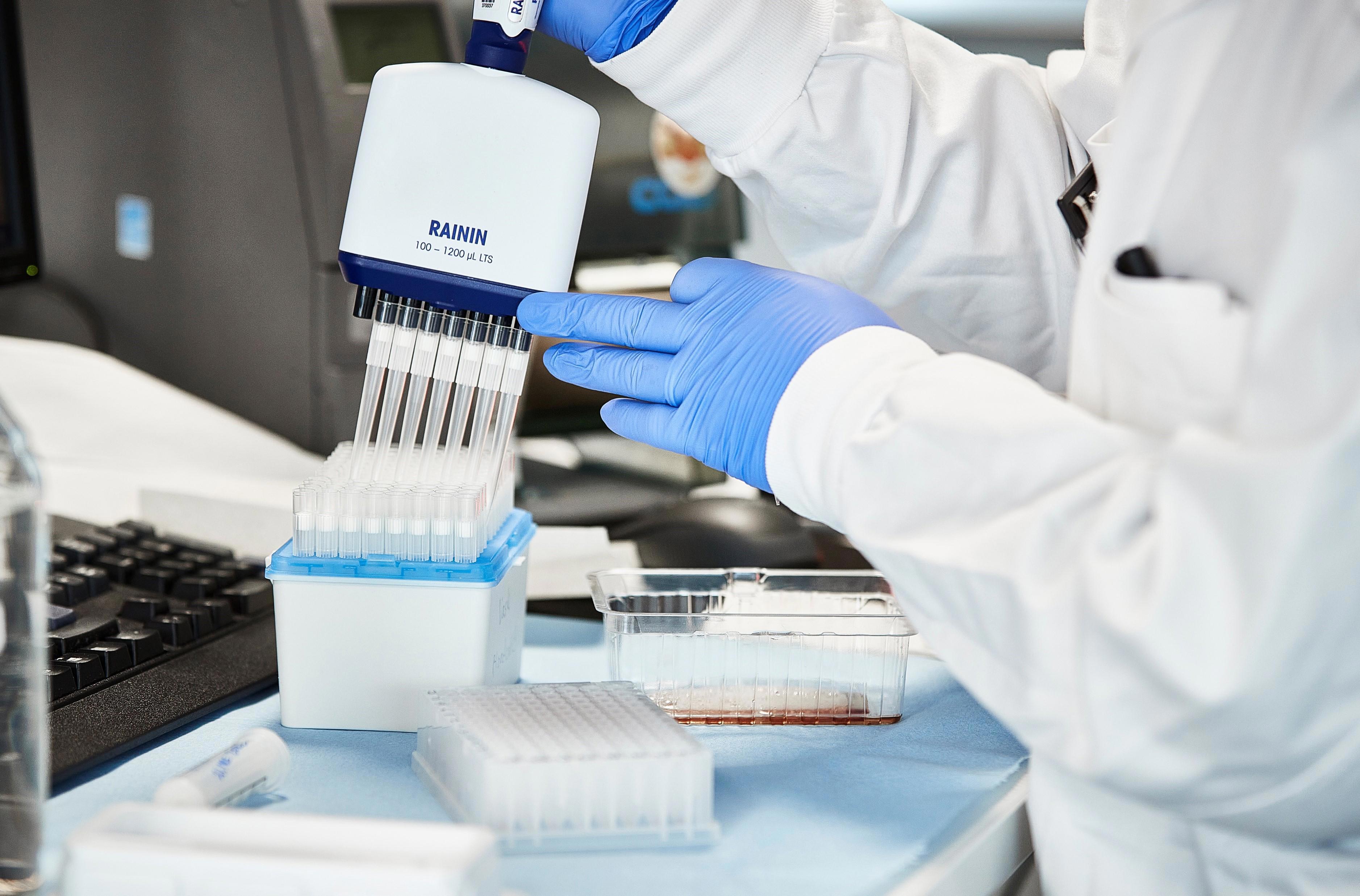 A laboratory technician wearing a white lab coat and blue gloves is using a multi-channel pipette to transfer liquid into a row of test tubes held in a blue and white rack on a lab bench. Additional lab equipment and containers are visible in the background, including a computer monitor and keyboard.