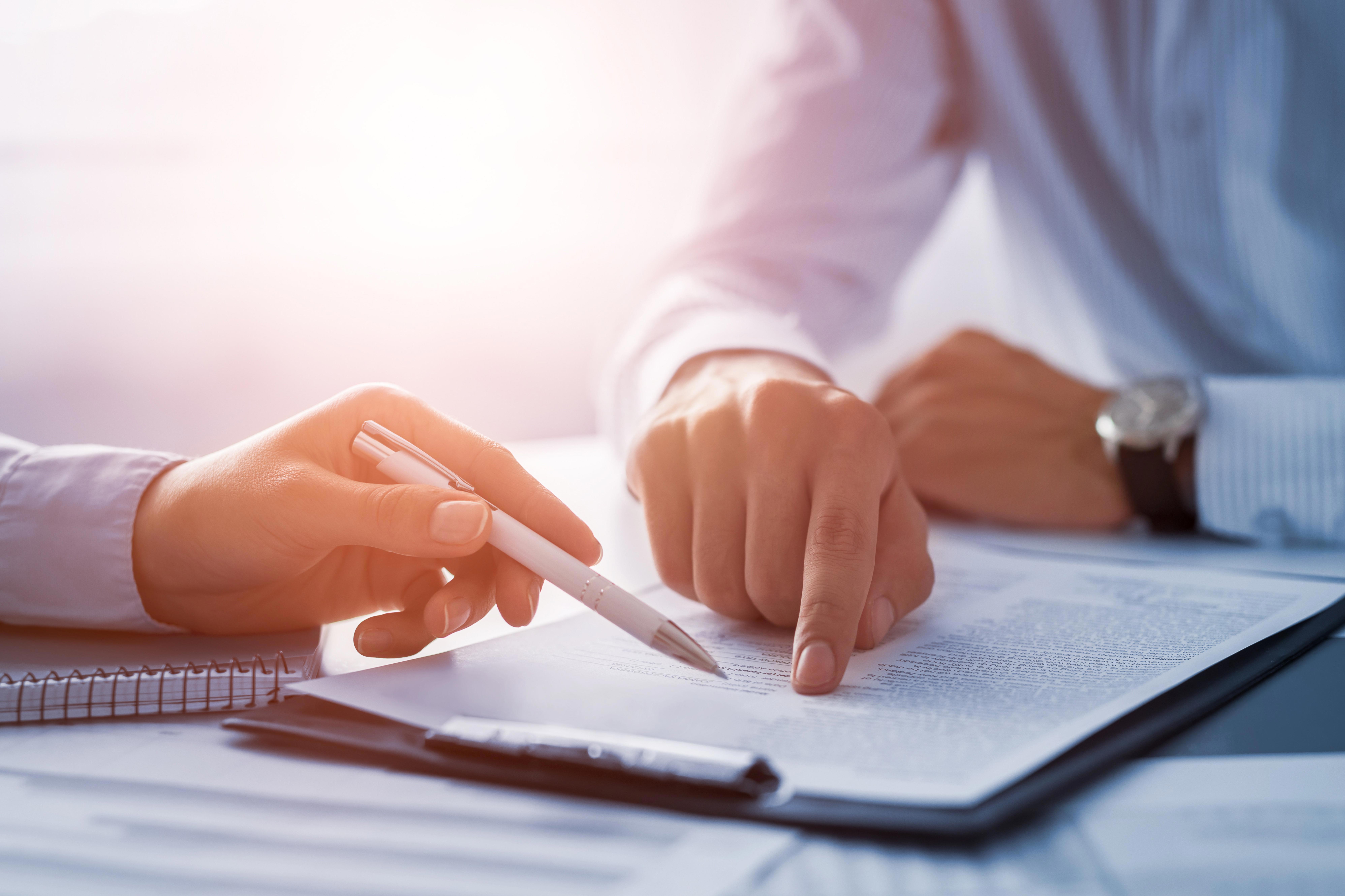 Two people are sitting at a table, reviewing and discussing a document. One person is holding a pen and pointing to the document, while the other person's hand is also visible, indicating collaboration or guidance.