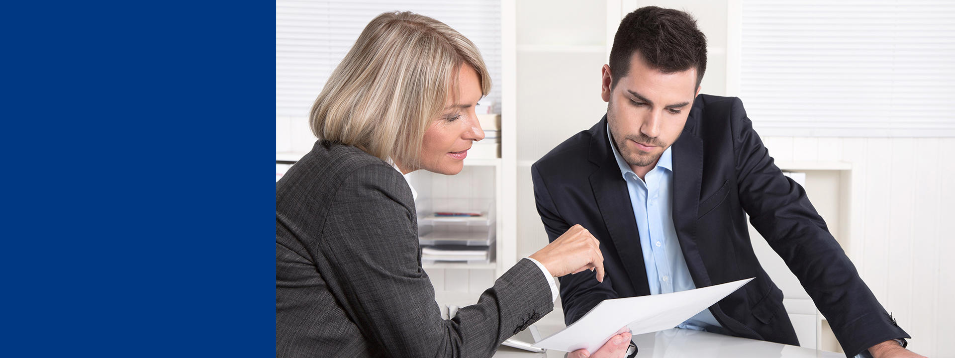 Two professionals in business attire seated at a table, collaborating while reviewing a printed document together, with one pointing to specific details on the page during discussion.