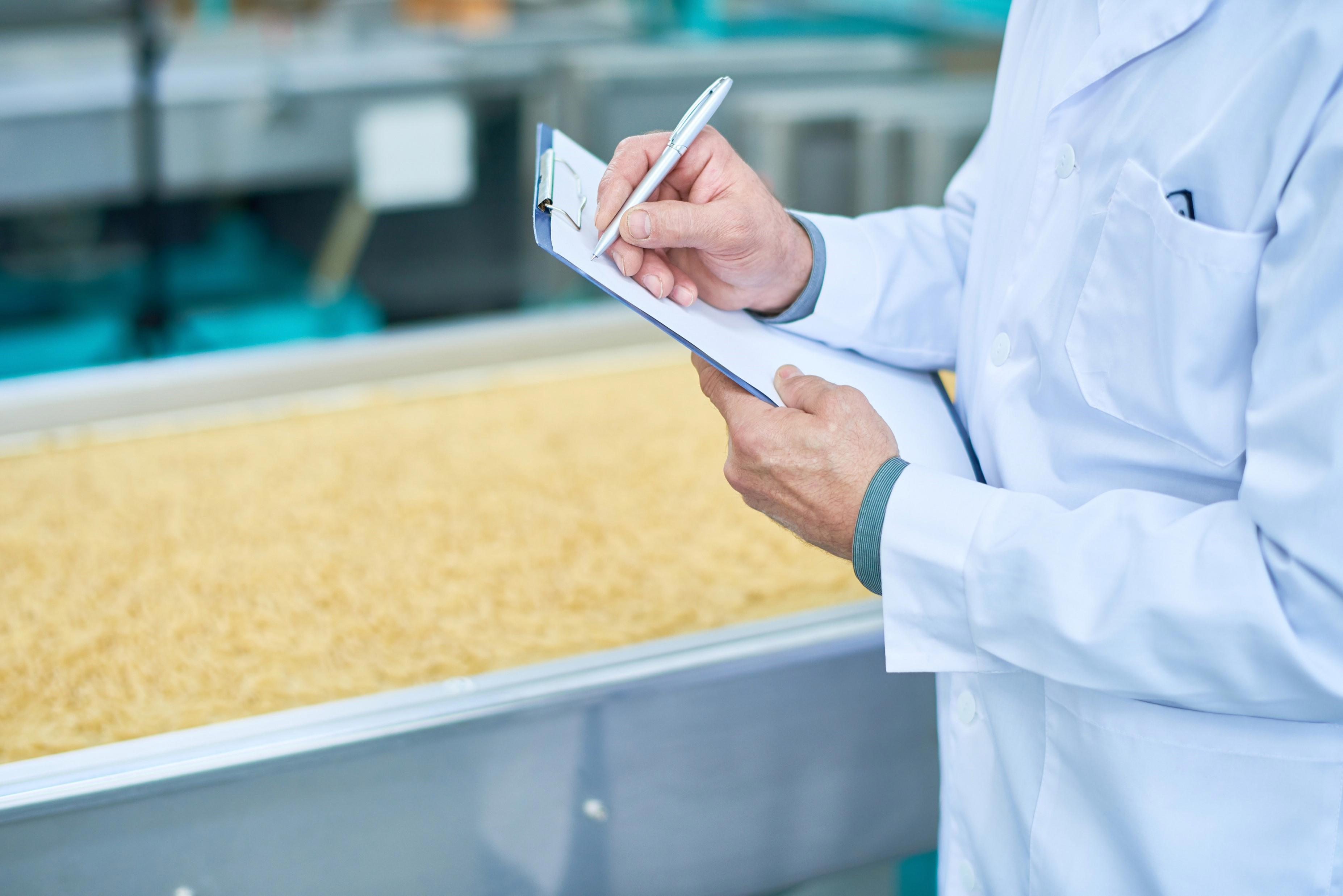 A person in a white lab coat is holding a clipboard and pen, writing or taking notes. The background shows a food production line with a large quantity of pasta or similar product being processed. This image depicts quality control or inspection in a food manufacturing facility.