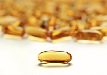 Close-up image of a single amber-colored softgel capsule containing a liquid or semi-liquid substance. Several similar capsules are visible in the blurred background.