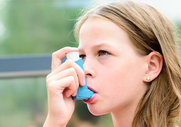 Image of a person using an inhaler. The hand holding the inhaler and part of the ear with an earring are visible. The background shows an outdoor setting with greenery and a distant structure.
