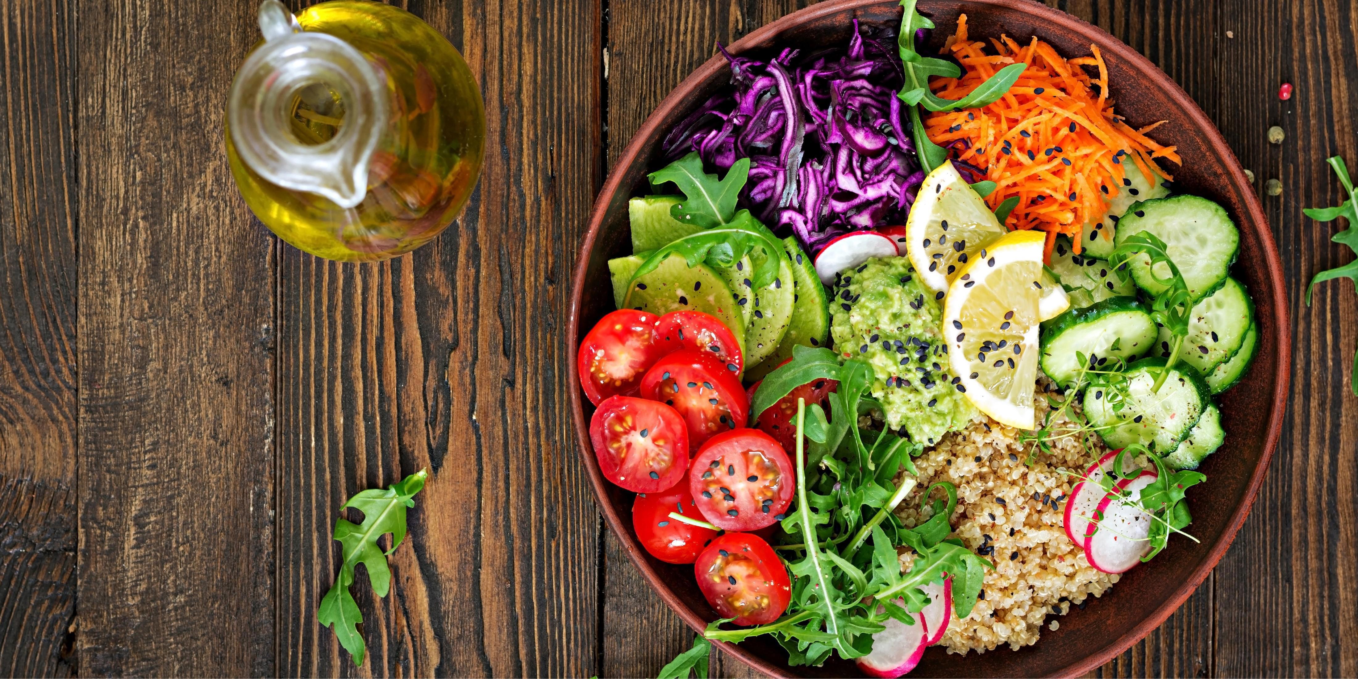 A wooden table with a bowl of colourful salad ingredients including sliced cucumbers, cherry tomatoes, radishes, shredded carrots, purple cabbage, quinoa, lime wedges, guacamole topped with black sesame seeds, and arugula leaves. An olive oil bottle is placed to the left of the bowl.
