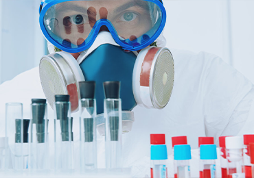 Person wearing full protective gear, including a respirator mask and goggles, working in a sterile laboratory environment. Various test tubes and vials with colored caps are arranged on the lab bench in front of them.