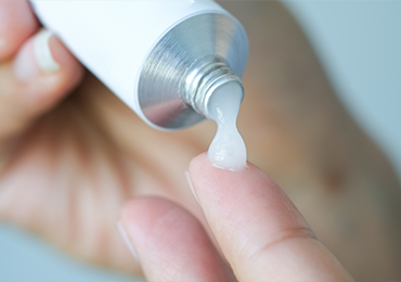 Close-up image of a person dispensing a semi-solid substance, such as an ointment or cream, from a metallic tube onto their fingertip. The focus is on the semi-solid dosage form being released from the tube