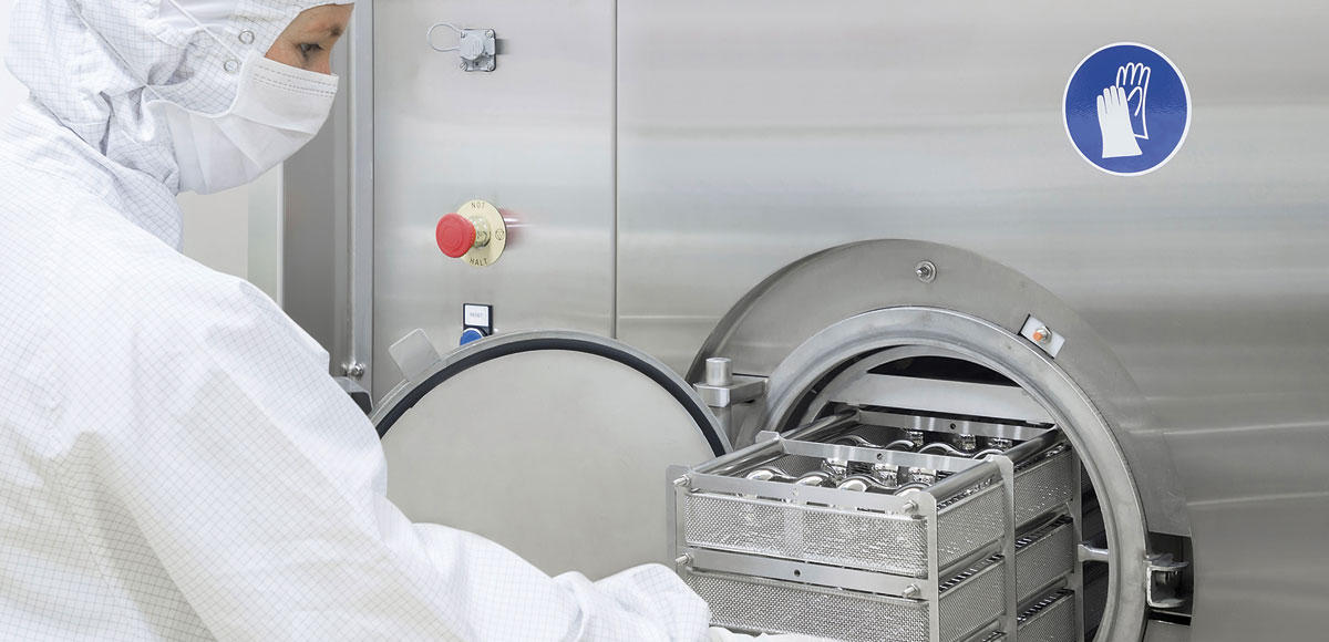 Person in Schutzkleidung bedient eine große industrielle Maschine aus Edelstahl mit geöffneter runder Tür, die Tabletts oder Behälter im Inneren zeigt. Die Maschine verfügt über einen roten Not-Aus-Schalter und ein blaues Sicherheitsschild mit Handsymbol.