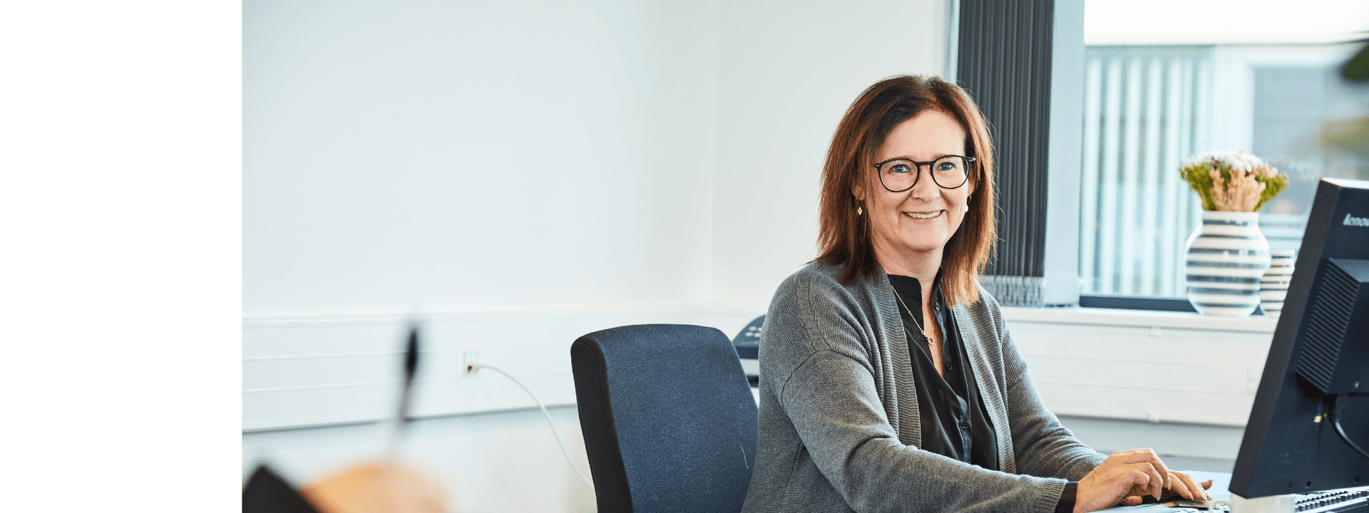 Mitarbeiterin mit Brille arbeitet lächelnd an einem Computerarbeitsplatz im Büro; im Hintergrund sind ein Fenster mit Jalousien, ein Blumenstrauß und Büroausstattung zu sehen.