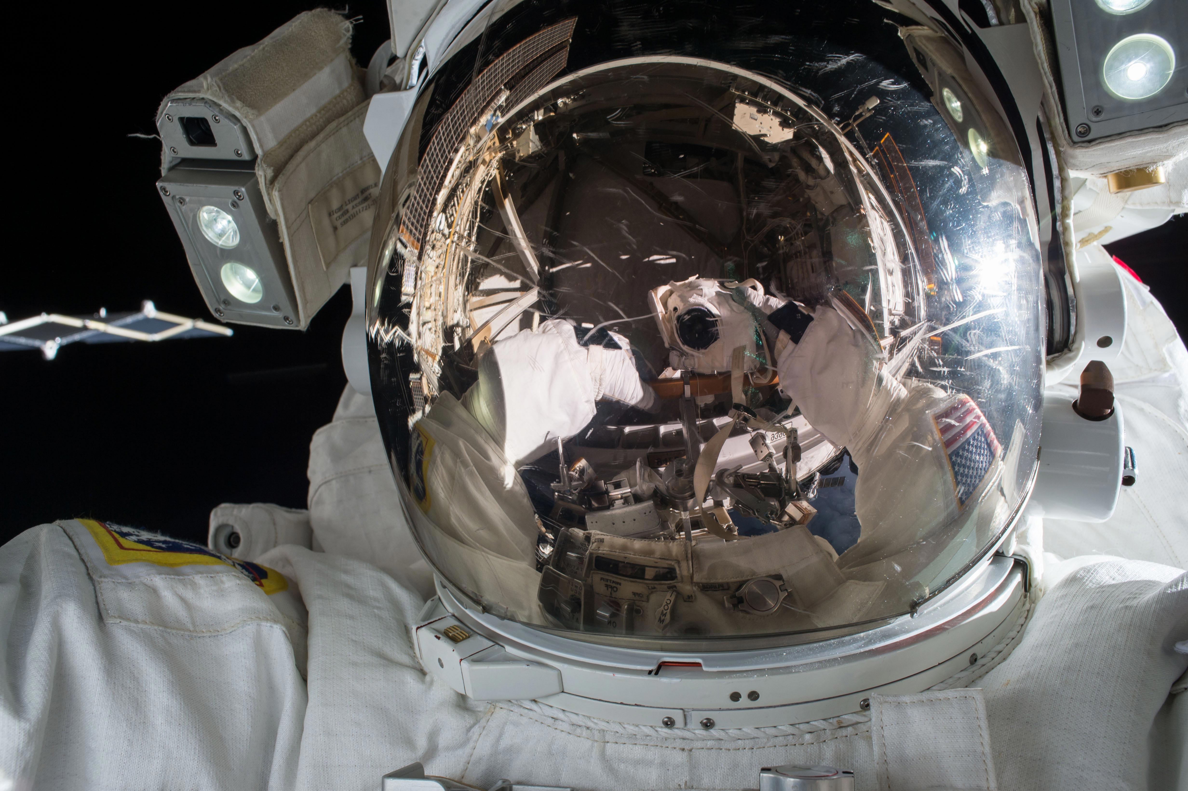 A close-up image of an astronaut in a spacesuit during a spacewalk. The astronaut's helmet visor reflects the intricate details of the spacecraft and another astronaut taking a photograph. The background is the dark expanse of space, with some parts of the spacecraft visible.
