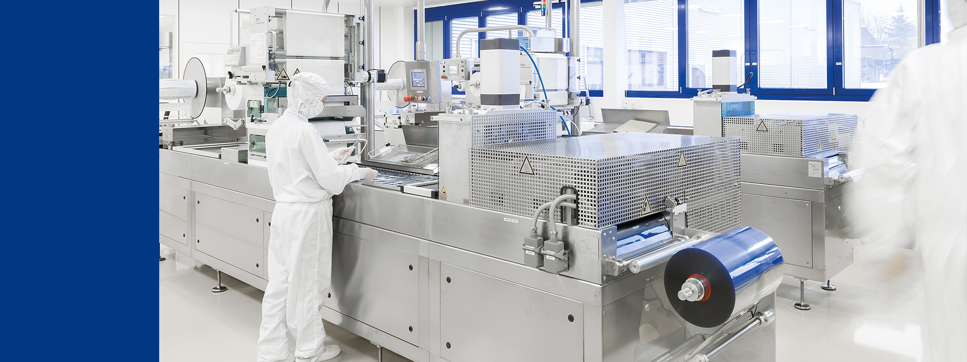 Mehrere Mitarbeiterinnen und Mitarbeiter in vollständiger Reinraumschutzkleidung bedienen und überwachen große Verpackungsmaschinen in einer Reinraum-Produktionsumgebung. 