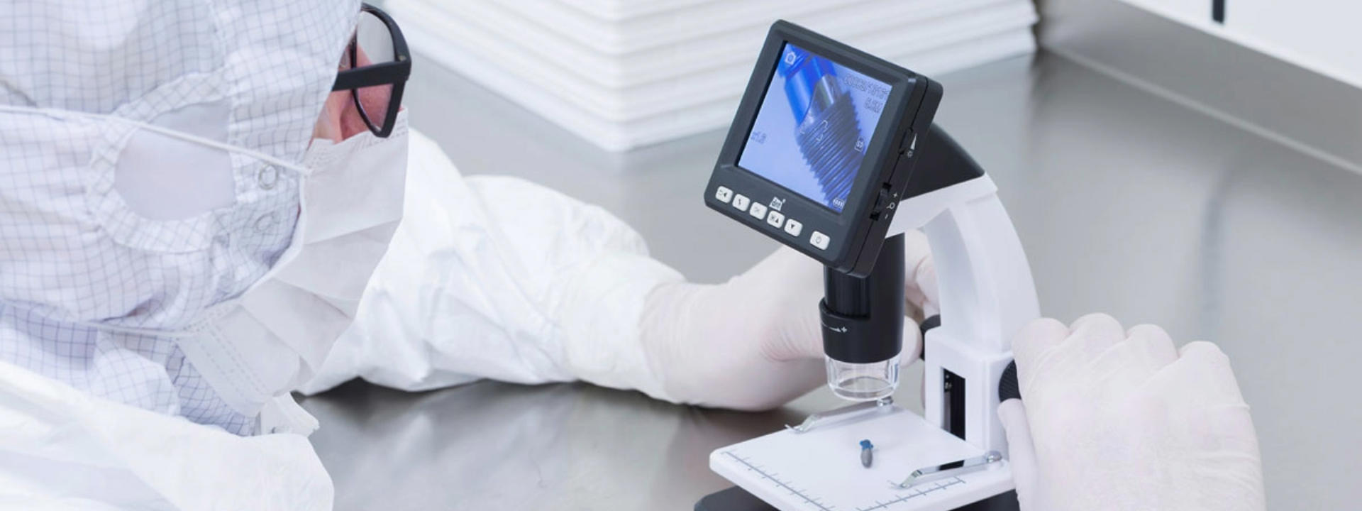 Fachkraft in Schutzkleidung bei der mikroskopischen Analyse mit einem digitalen Mikroskop in einer Reinraumumgebung. Auf dem Bildschirm ist eine vergrößerte Darstellung der Probe zu sehen