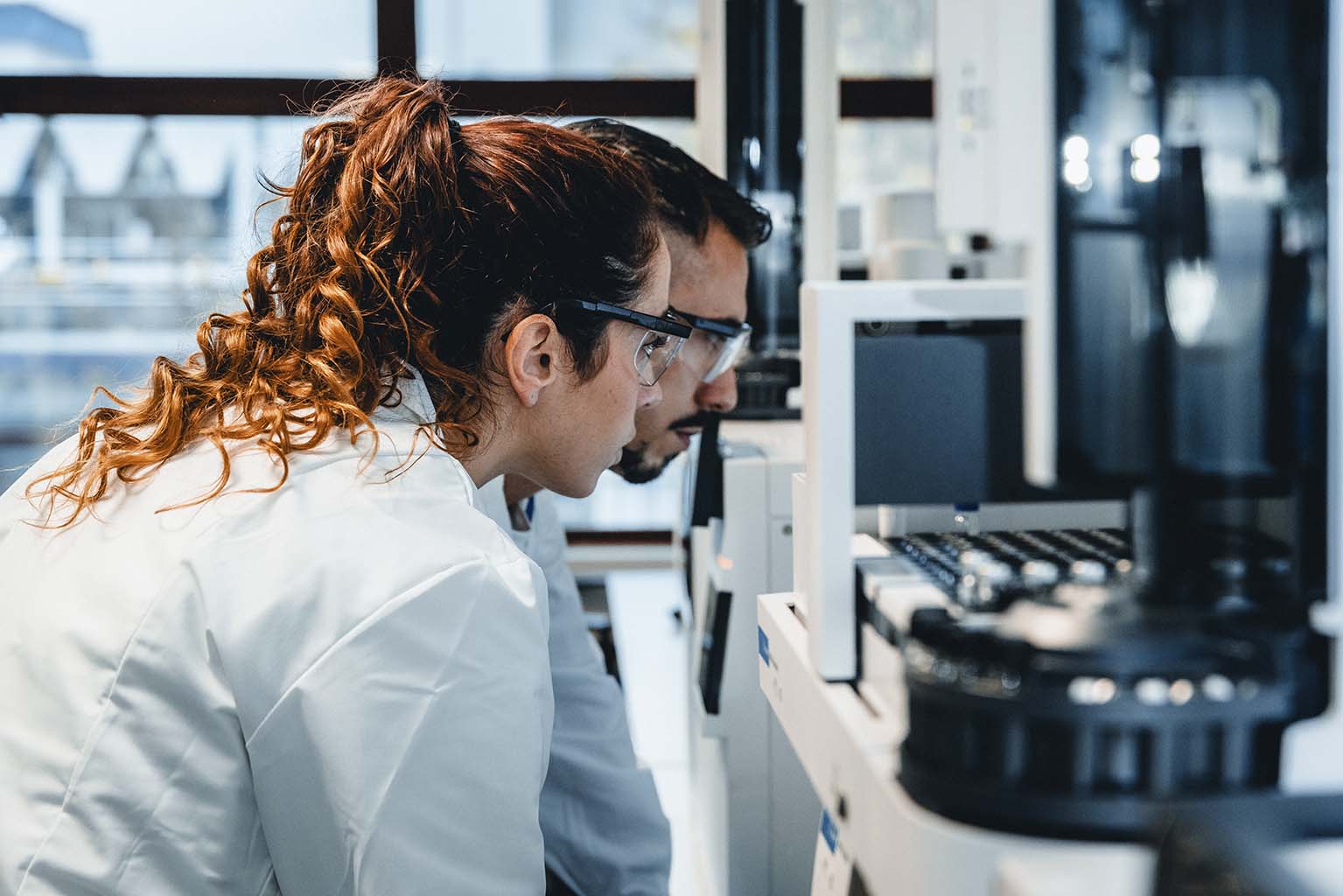 Zwei Fachkräfte in Laborkitteln und Schutzbrillen analysieren konzentriert medizinisches Equipment oder Daten auf einem Bildschirm in einem modernen Labor. Im Hintergrund sind Laborgeräte sichtbar.