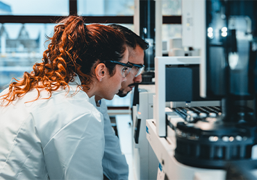 Zwei Fachkräfte in Laborkitteln und Schutzbrillen analysieren konzentriert medizinisches Equipment oder Daten auf einem Bildschirm in einem modernen Labor. Im Hintergrund sind Laborgeräte sichtbar.