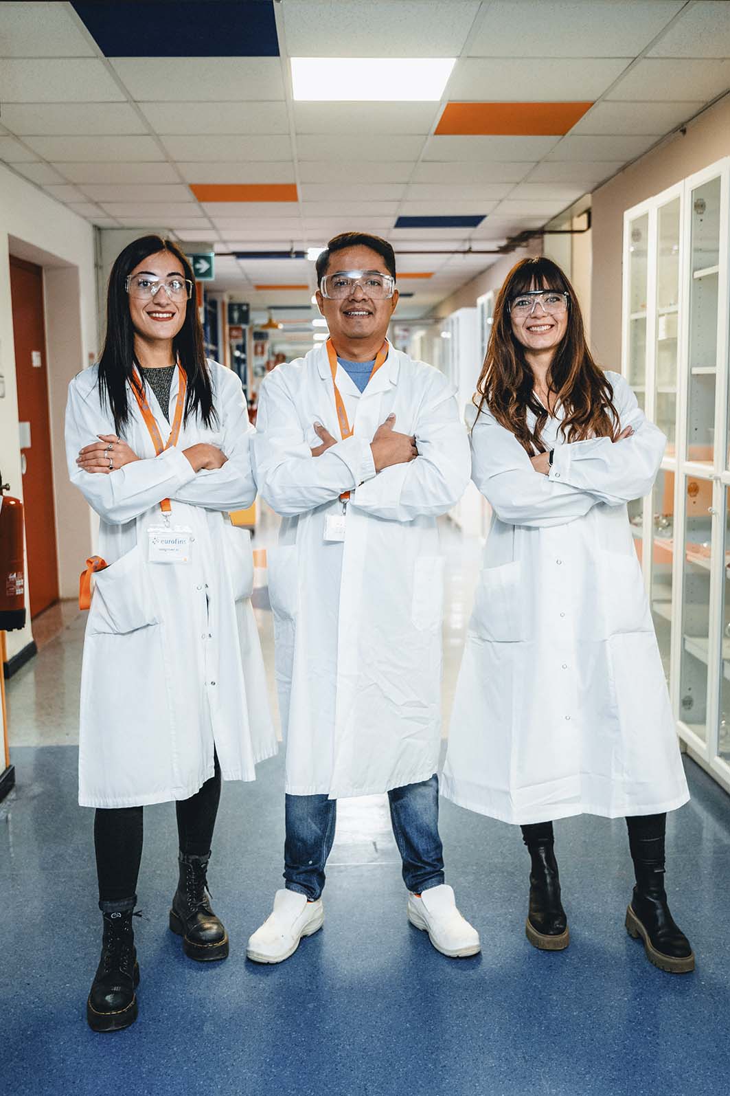 Drei Fachkräfte in weißen Laborkitteln und Schutzbrillen stehen mit verschränkten Armen in einem farblich gestalteten Flur eines Labors.
