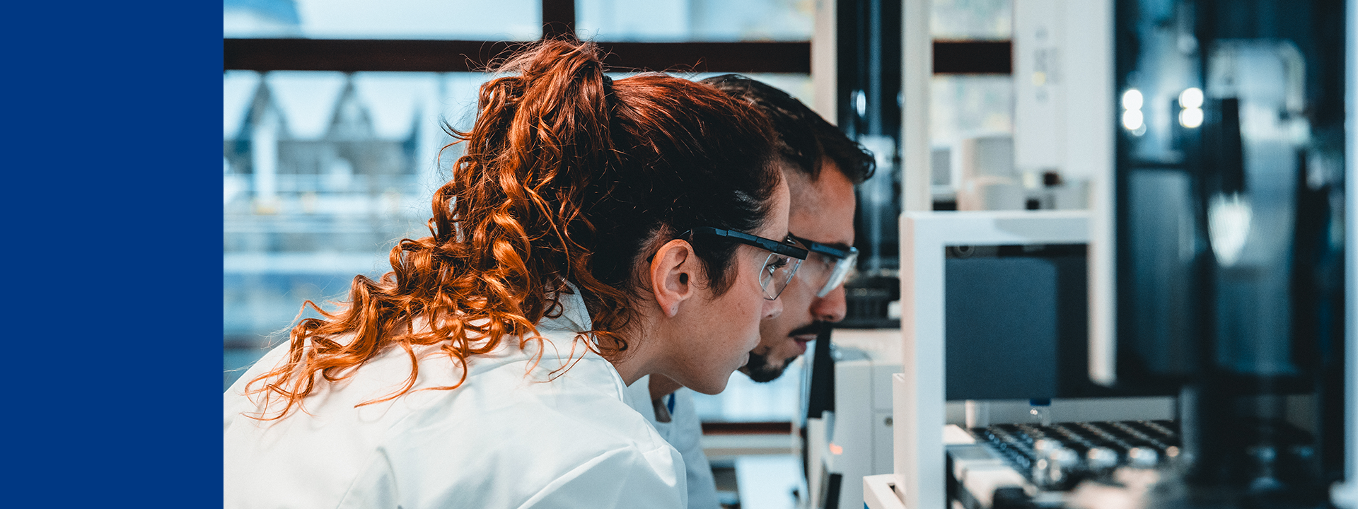 Zwei Fachkräfte in Laborkitteln und Schutzbrillen analysieren konzentriert medizinisches Equipment oder Daten auf einem Bildschirm in einem modernen Labor. Im Hintergrund sind Laborgeräte sichtbar.