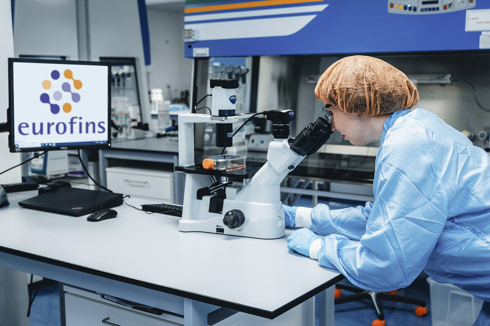 Person in a blue lab coat and orange hairnet looking through a microscope in a laboratory. A computer monitor on the left displays the Eurofins logo and text. Various scientific instruments are visible in the background