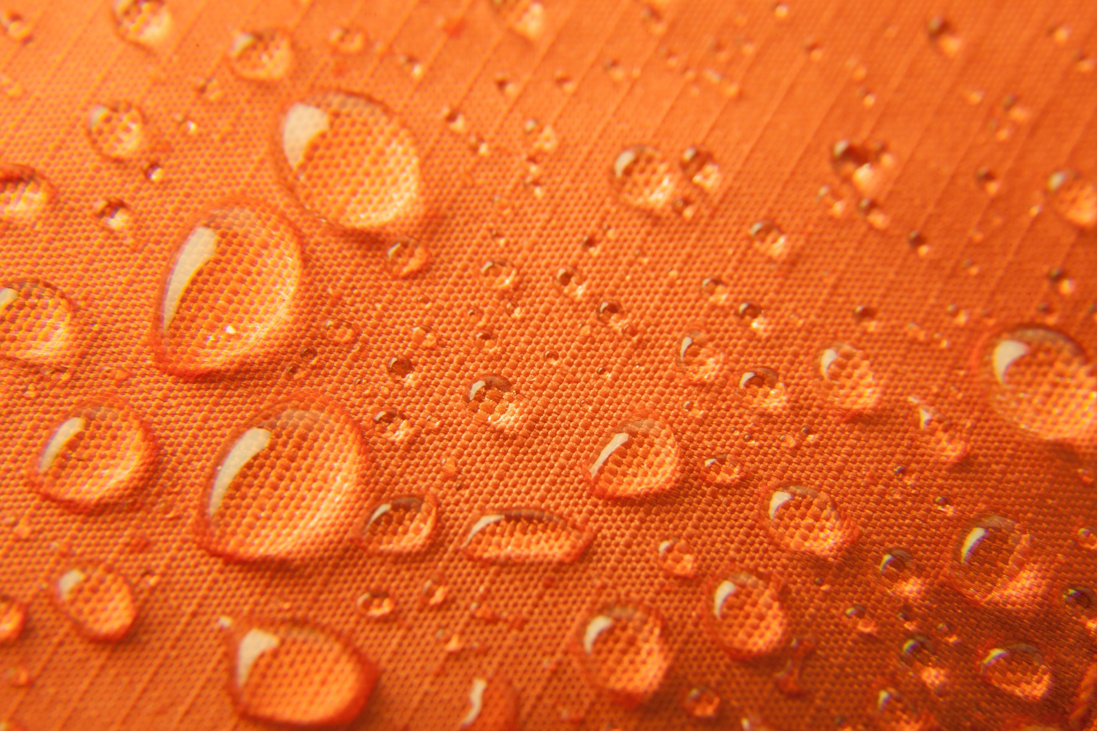 Water droplets on an orange, textured surface. The surface has a grid-like pattern, and the droplets vary in size, creating a visually appealing contrast between the smooth water and the rough texture of the fabric.
