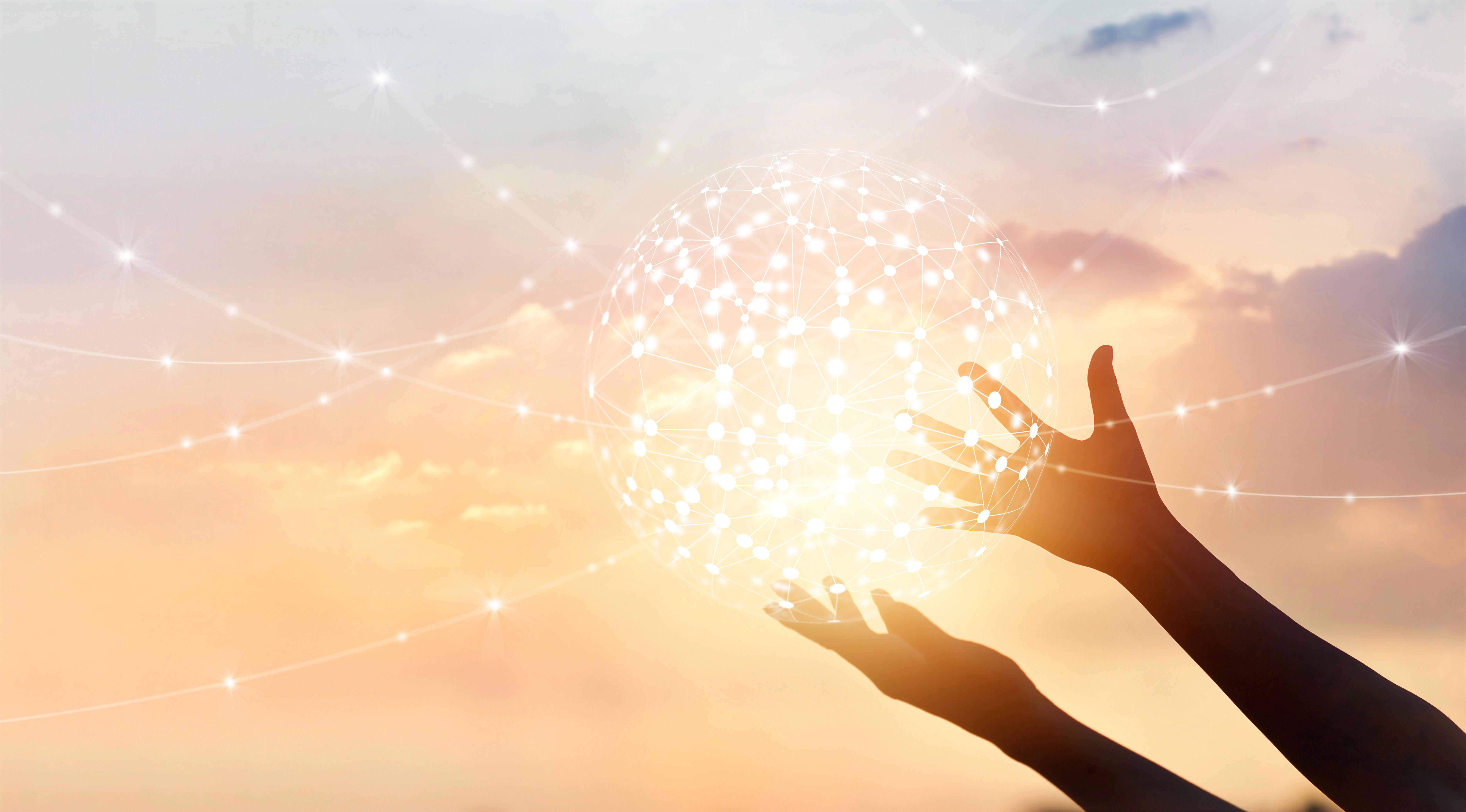  Two hands are reaching up towards a glowing, interconnected sphere of light against a soft, pastel-coloured sky at sunset. The sphere is composed of numerous points connected by lines, resembling a network or constellation. 