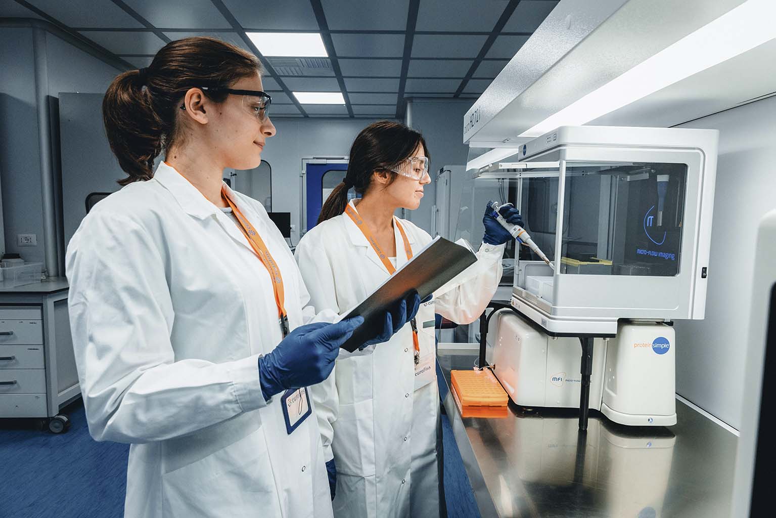 Zwei Fachkräfte in weißen Laborkitteln, Schutzbrillen und blauen Handschuhen arbeiten in einem Labor. Eine Person bedient eine Pipette an einem Laborgerät, während die andere mit einem Klemmbrett beobachtet.