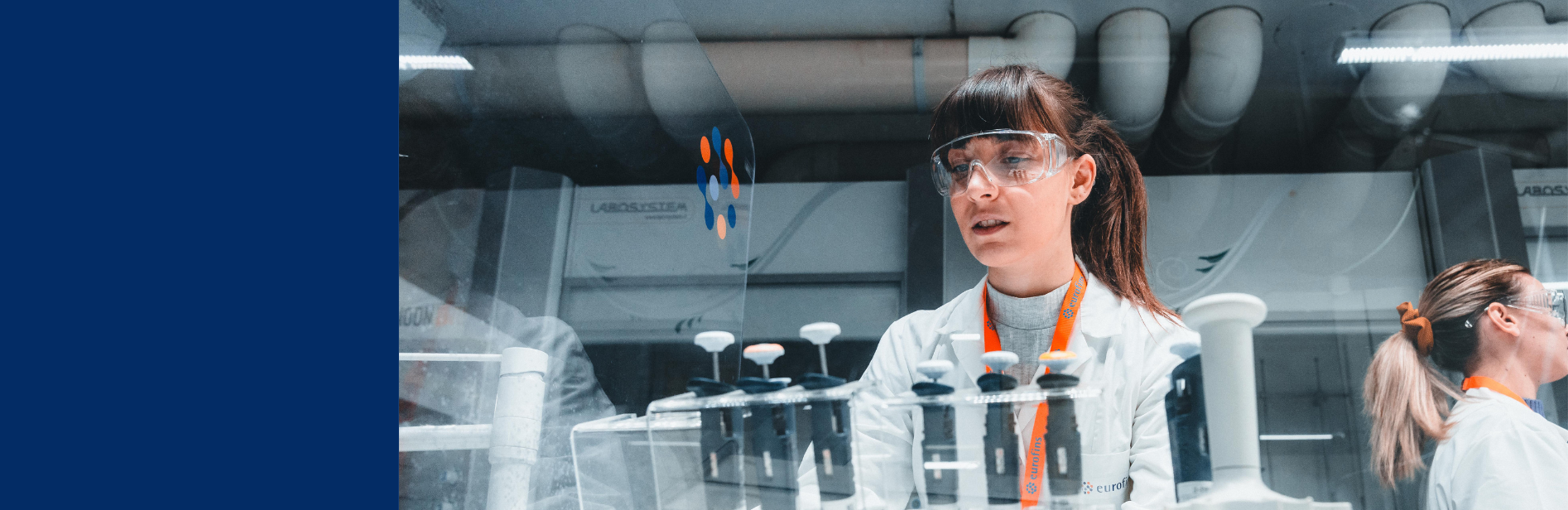 Wissenschaftlerin im Labor mit Schutzbrille und Laborkittel, arbeitet mit Pipetten und Reagenzgläsern inmitten moderner Forschungsausrüstung und industrieller Maschinen.