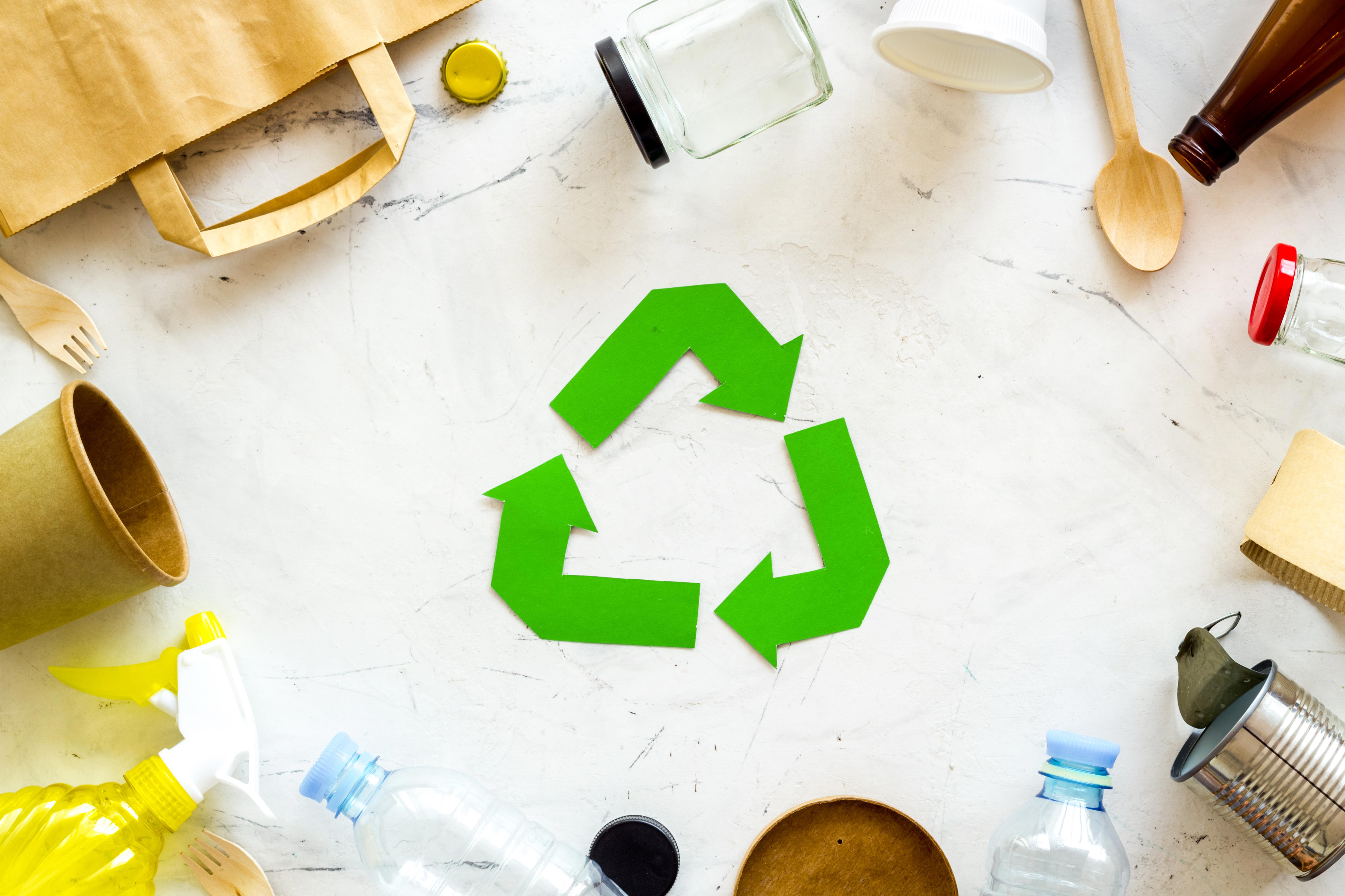 ecology-concept-green-recycling-sign-with-waste-materials-paper-bag-and-cup-plastic-bottles-flatware-on-white-background-top-view_7278-x-4852-px