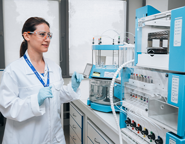 Laborfachkraft mit Schutzbrille und Handschuhen bedient ein komplexes Laborgerät mit mehreren Schläuchen und Behältern.
