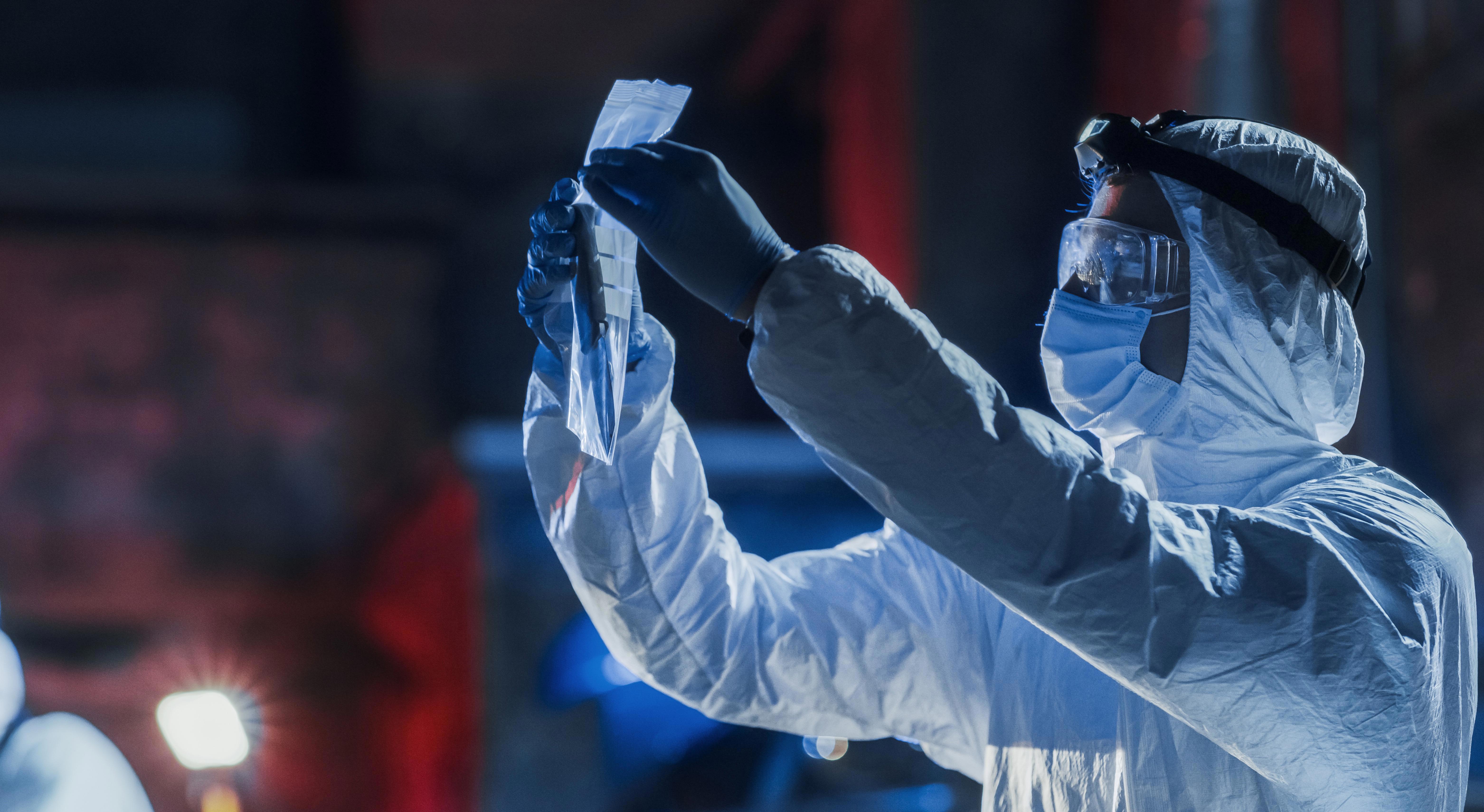 A forensic investigator in a full protective suit examining evidence at a crime scene. The person is holding a plastic bag, examining its contents..