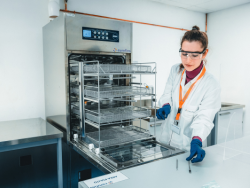 Laborfachkraft mit Schutzbrille und Handschuhen bei der Aufbereitung und Inspektion von metallischen Medizinprodukte-Komponenten an einem geöffneten Sterilisator.