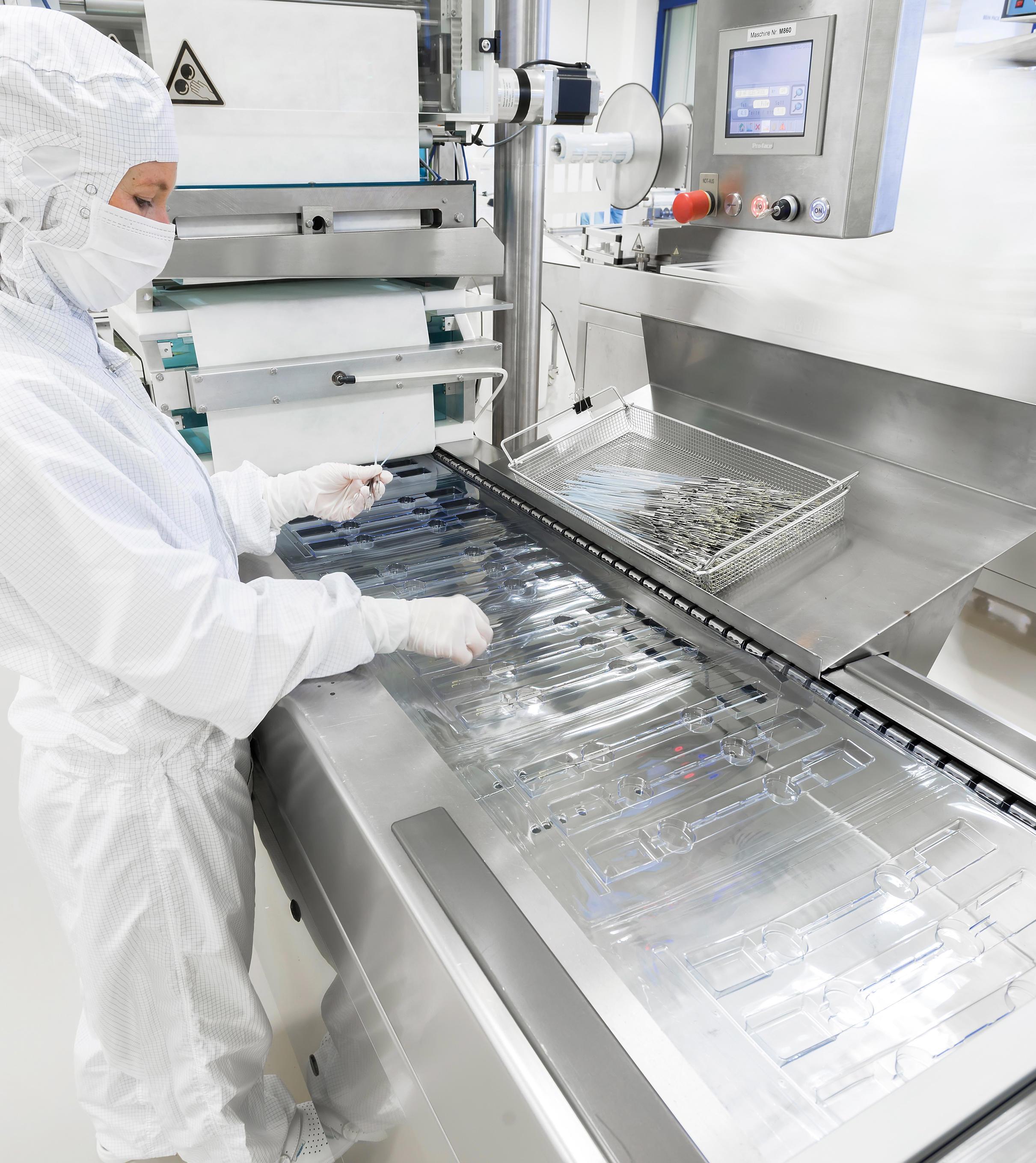 Person in Schutzkleidung arbeitet in einer Reinraumumgebung, hantiert mit transparenter Kunststoffverpackung auf einem Förderband, umgeben von Fertigungsanlagen und Steuerungspaneelen.