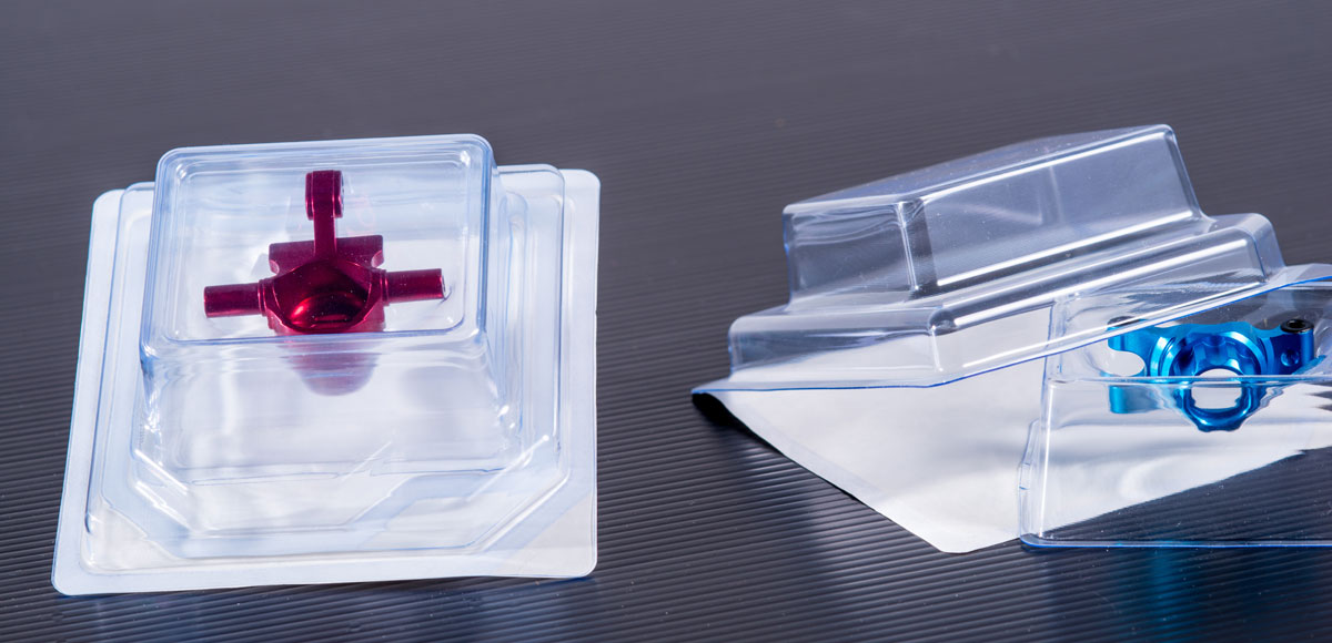 Zwei transparente Kunststoffverpackungsbehälter, einer mit einem roten metallischen Objekt und der andere mit einem blauen metallischen Objekt, auf einer dunklen Oberfläche platziert.