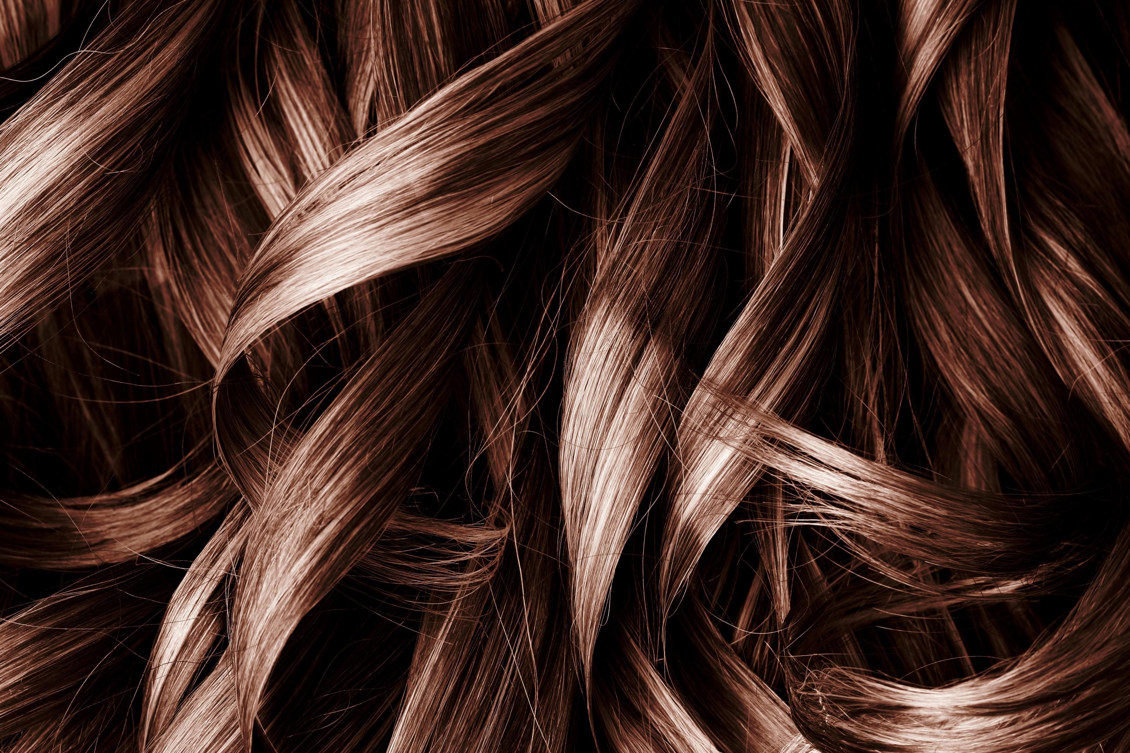 A close-up image of wavy, shiny, brown hair strands intertwined and overlapping each other. The texture and sheen of the hair are clearly visible, highlighting the smoothness and healthiness of the hair.