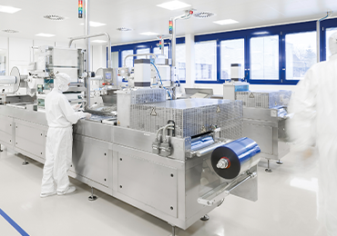 Mehrere Mitarbeiterinnen und Mitarbeiter in vollständiger Reinraumschutzkleidung bedienen und überwachen große Verpackungsmaschinen in einer Reinraum-Produktionsumgebung. 