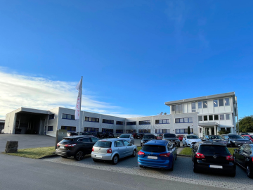 Modernes Bürogebäude mit mehreren Fenstern und Flachdach, davor ein Parkplatz mit verschiedenen Autos. Der Himmel ist klar und blau, und in der Nähe des Eingangs steht ein Fahnenmast mit einer Flagge.