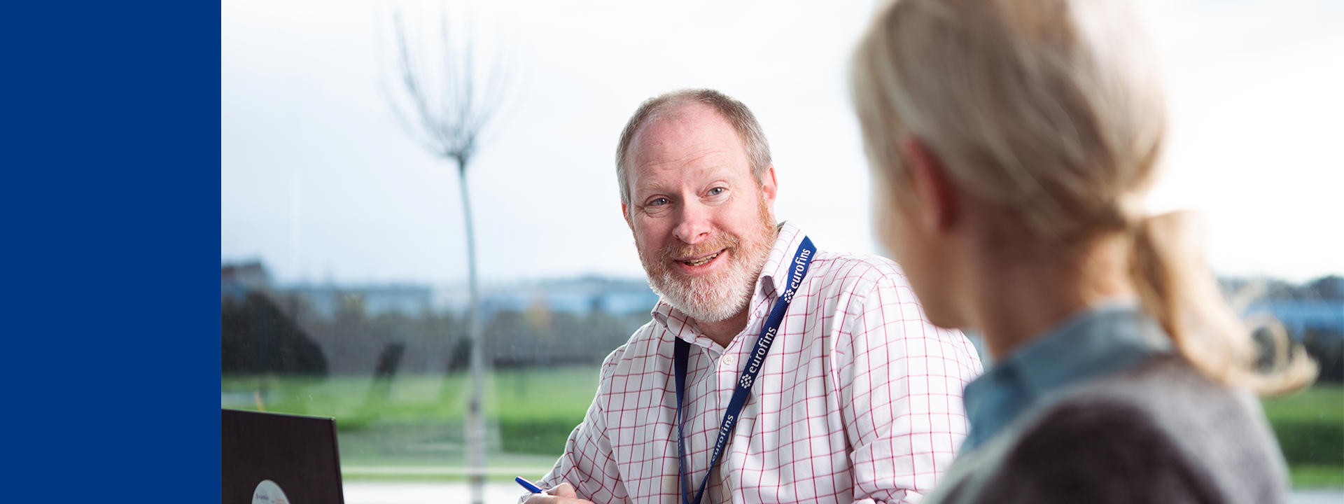 Zwei Personen im Gespräch in einem modernen Innenraum mit großen Fenstern und Blick ins Grüne. Eine Person trägt ein Namensschild.