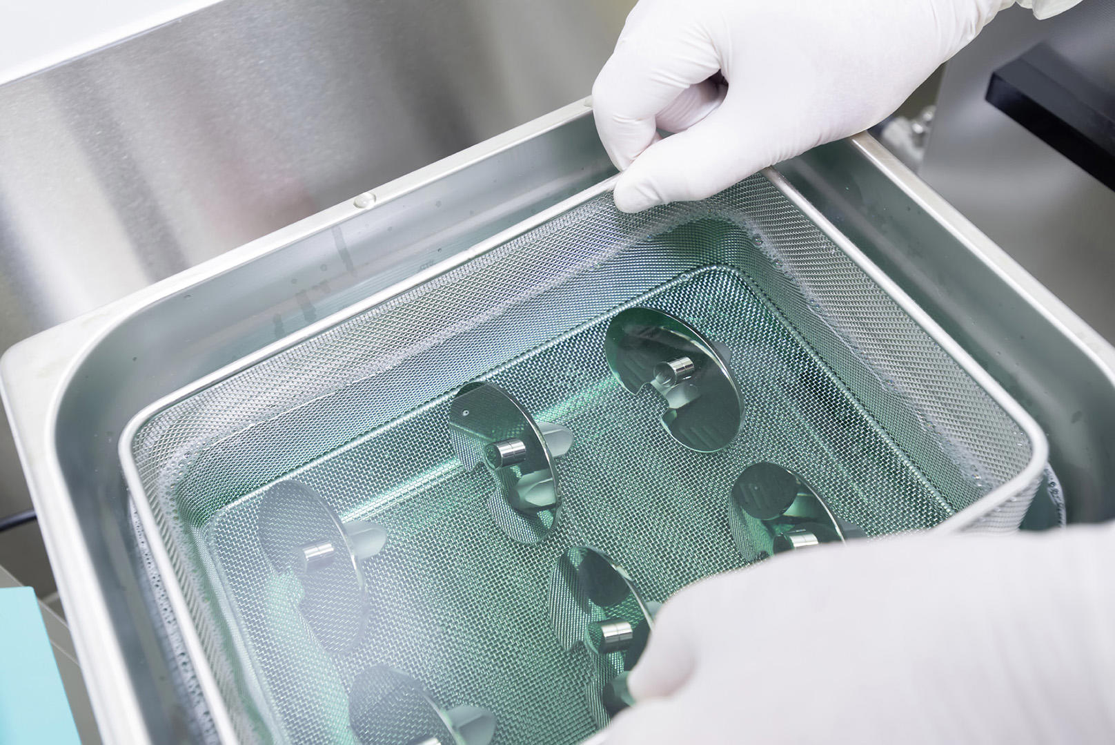 Behandschuhte Hände platzieren einen Metallkorb mit mehreren Medizinprodukten in einen Edelstahlbehälter. 