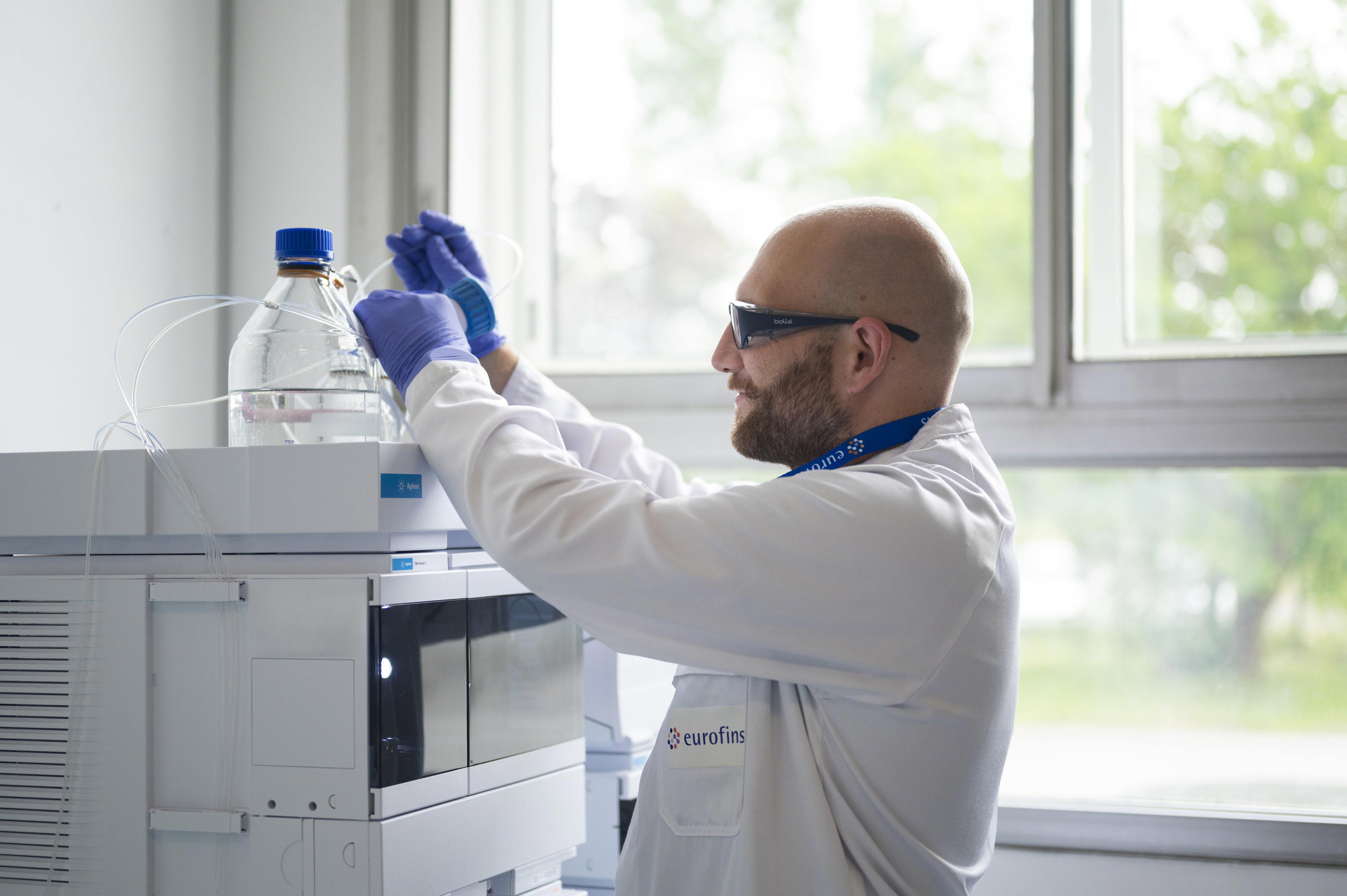 Scientifique Eurofins BioPharma Product Testing ajustant un dispositif de laboratoire pour le traitement d’échantillons liquides, illustrant l’application des lignes directrices ICH Q2(R2) et Q14 dans un environnement pharmaceutique contrôlé.