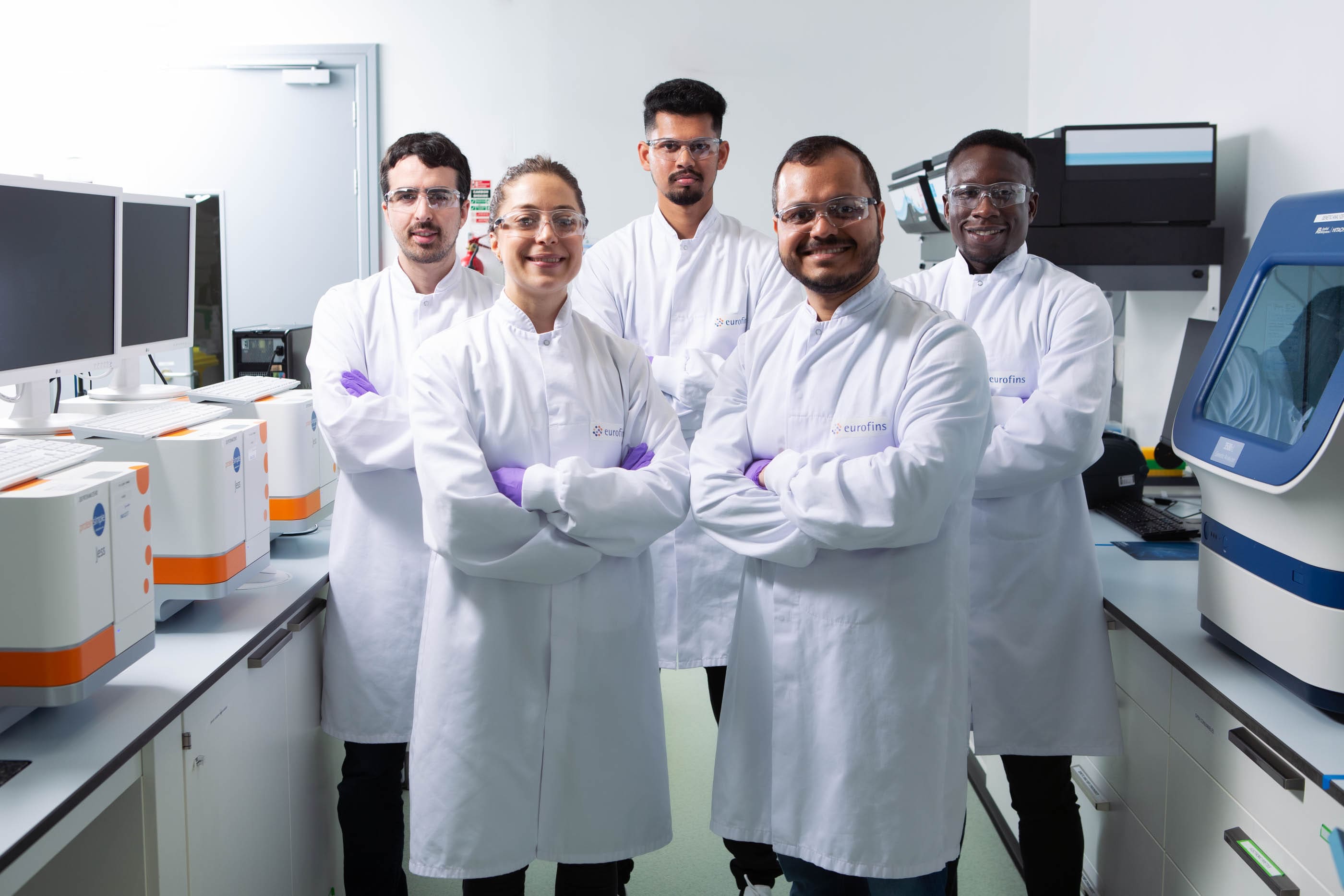 Cinq scientifiques d’Eurofins BioPharma Product Testing France en blouse blanche, lunettes de protection et gants, réunis dans un laboratoire moderne équipé d’ordinateurs et d’appareils analytiques. L’image illustre les activités de recherche collaborative et d’analyse des causes profondes dans un environnement conforme aux normes de qualité et de sécurité biopharmaceutiques.