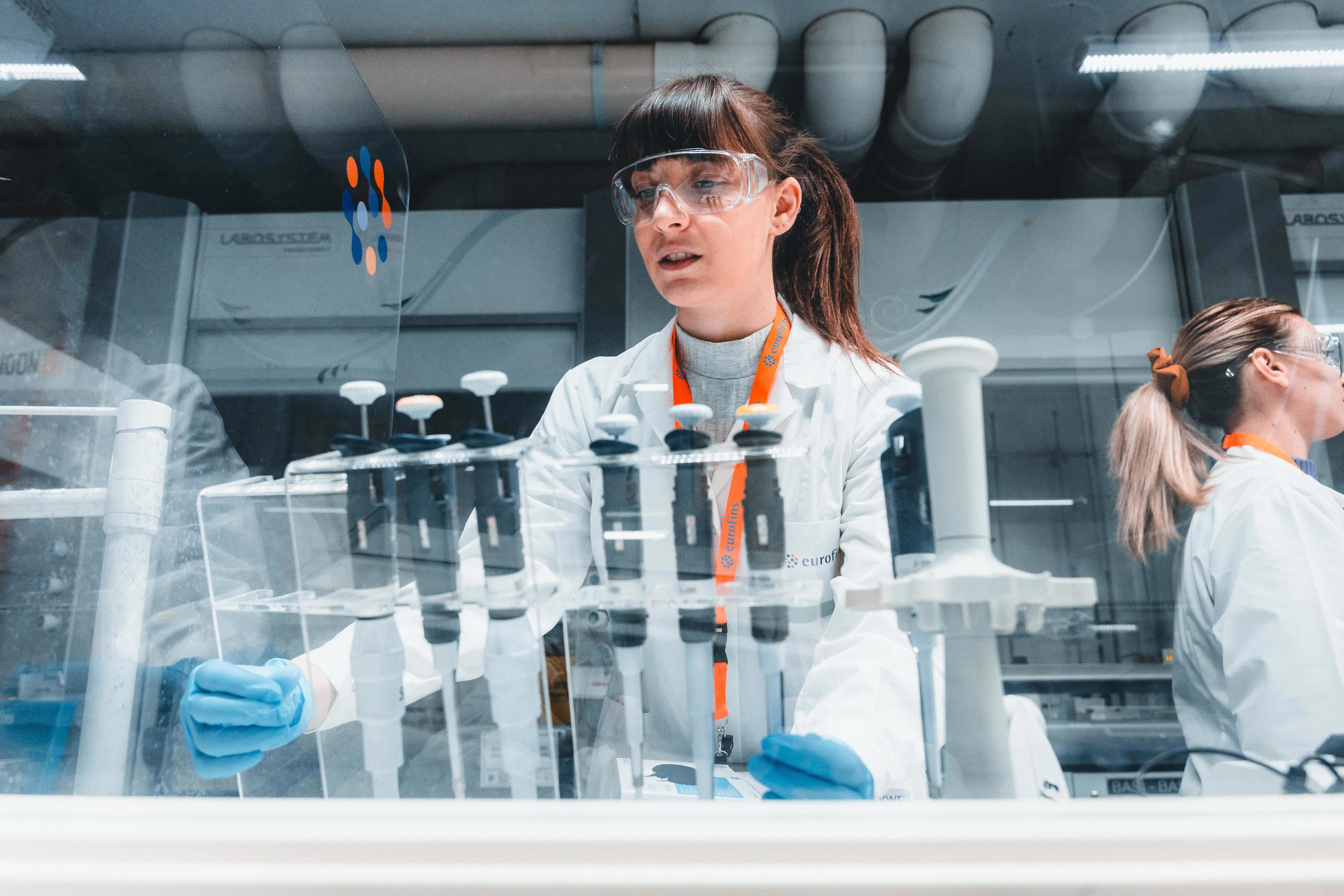 A laboratory scene showing two individuals wearing white lab coats and blue gloves working with scientific equipment. The person in the foreground is handling a rack of test tubes, while the person in the background is also engaged in laboratory work. The environment appears to be a modern, well-equipped lab with various instruments and tools visible.