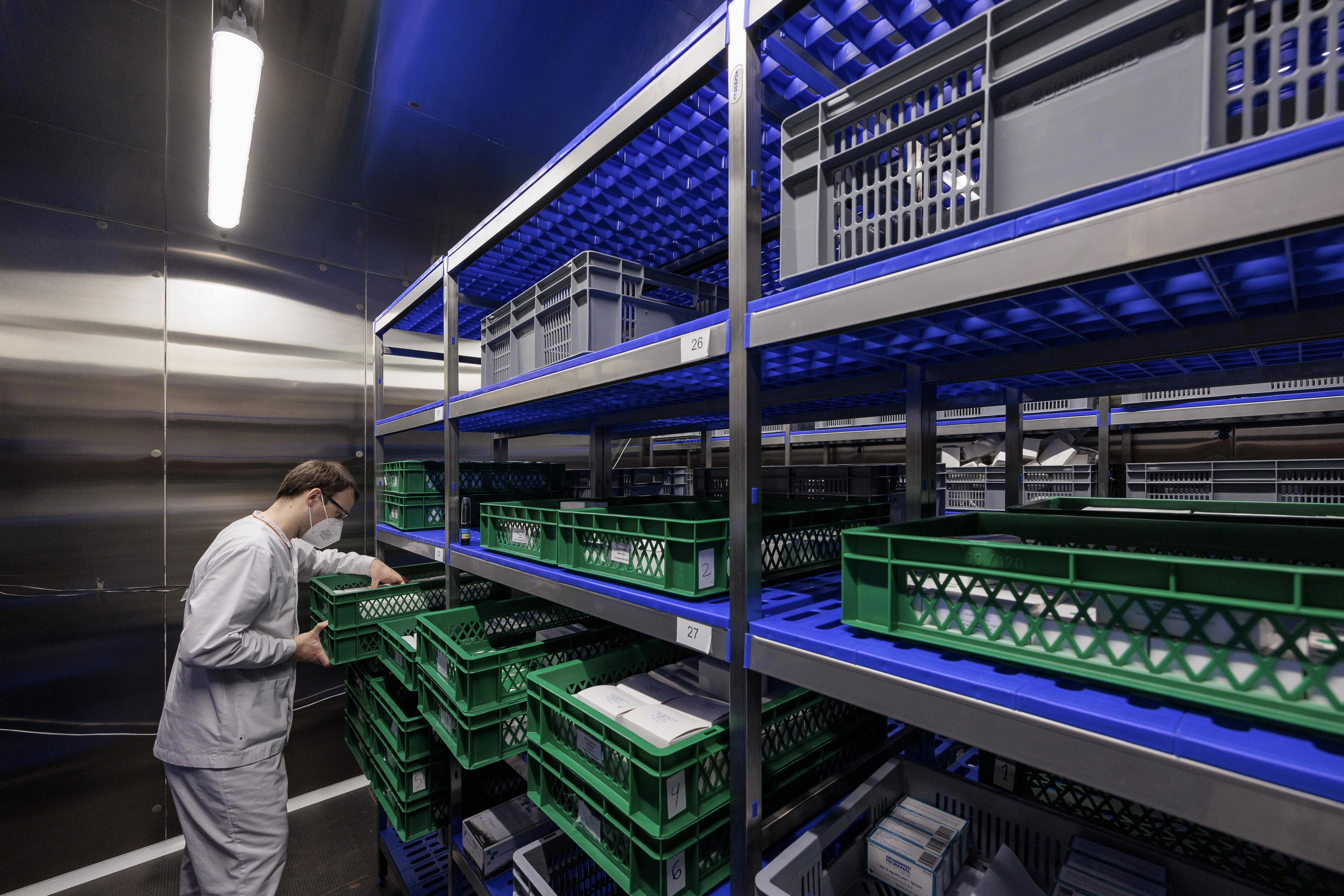 Collaborateur d’Eurofins BioPharma Product Testing en tenue stérile, avec masque et lunettes de protection, debout devant des étagères métalliques contenant des bacs en plastique verts et gris dans un espace de stockage pharmaceutique éclairé par des néons