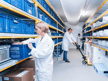 Techniciens en blouse blanche dans une zone de stockage pharmaceutique, manipulant des bacs et des palettes dans un entrepôt organisé. L’image illustre les solutions de stockage proposées par Eurofins Biopharma Product Testing pour les médicaments expérimentaux, incluant la gestion des stocks, le contrôle des températures (réfrigéré, congelé, ambiant), la traçabilité et le respect des bonnes pratiques de distribution (GDP) dans le cadre des essais cliniques.