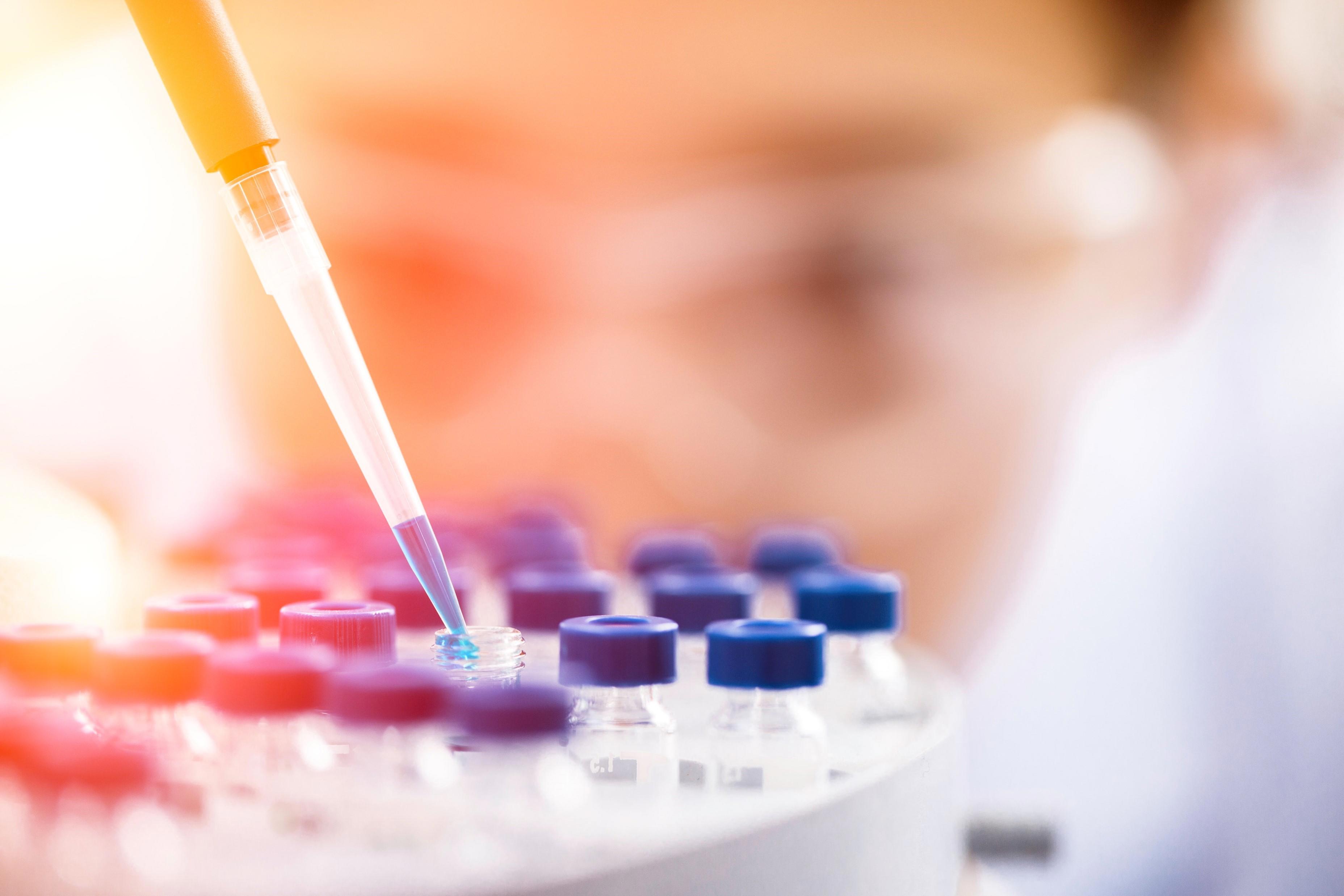A close-up image of a blue substance being pipetted into a vial. The vial is on a tray among other vials with blue lids. In the background, there is a blurred face wearing safety glasses.
