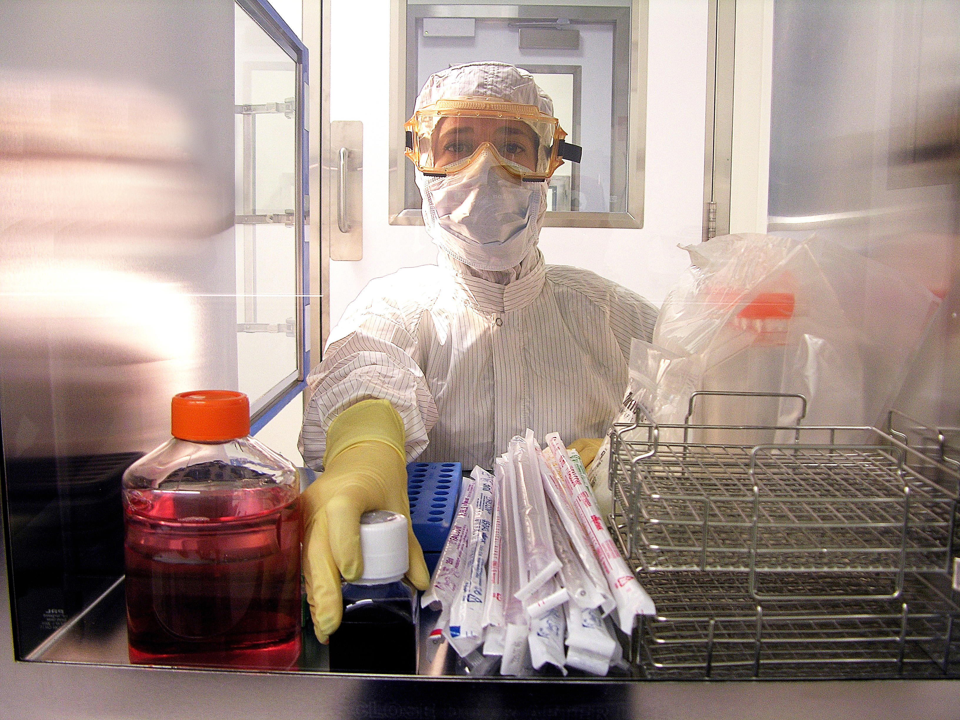 A person wearing protective gear, including a full-body suit, gloves, and a face mask, is working in a laboratory setting. The person is handling various lab equipment and supplies, including a container of red liquid on the left side, several individually wrapped pipettes in the centre, and metal racks on the right side. The background shows a cleanroom environment with stainless steel surfaces and glass windows.