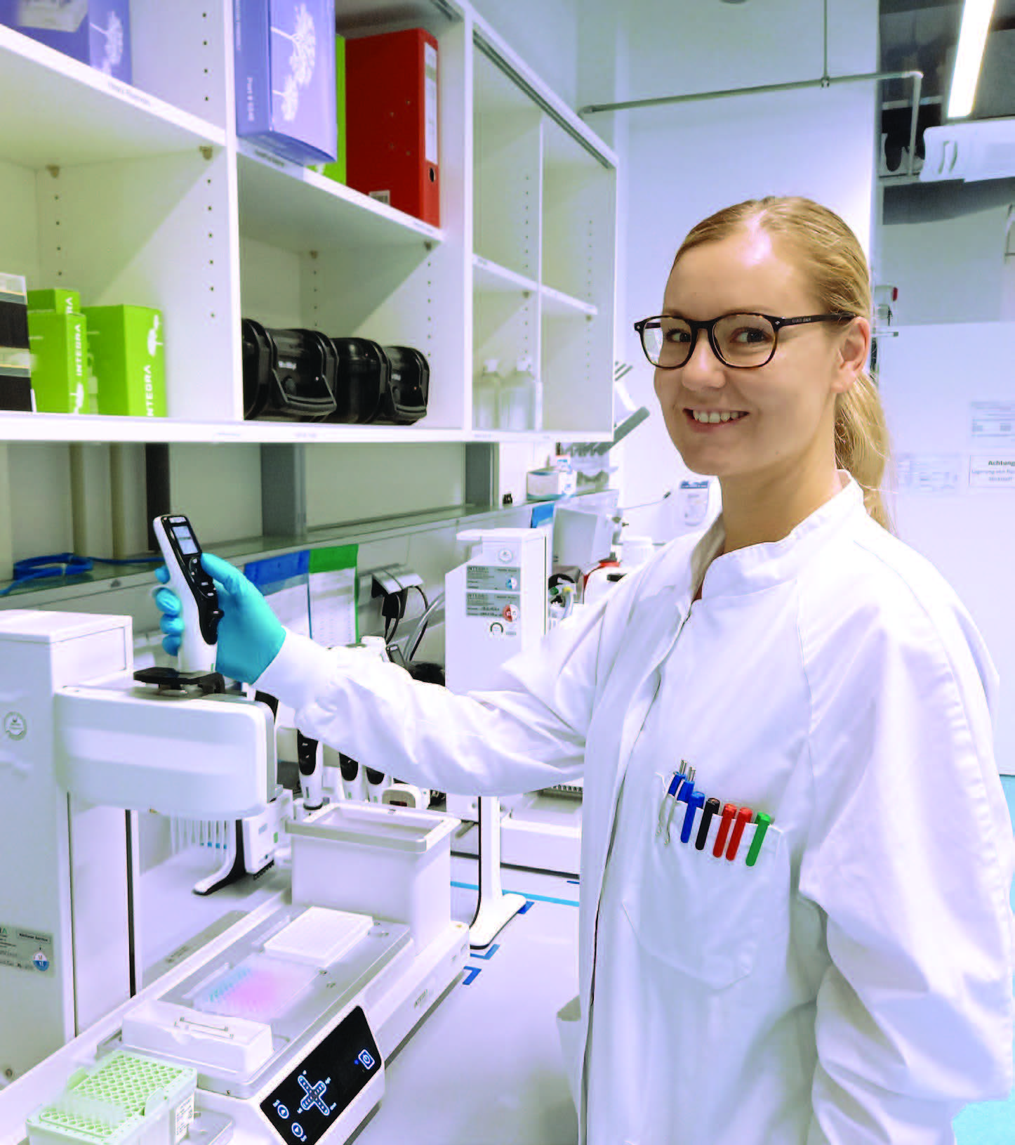 Personne en blouse blanche, lunettes de protection et gants bleus, manipulant un appareil électronique dans un laboratoire bien équipé. Elle se tient à côté d’un équipement automatisé, avec des boîtes et instruments scientifiques en arrière-plan. Cette scène illustre les activités de recherche ou de contrôle qualité menées par Eurofins BioPharma Product Testing France.