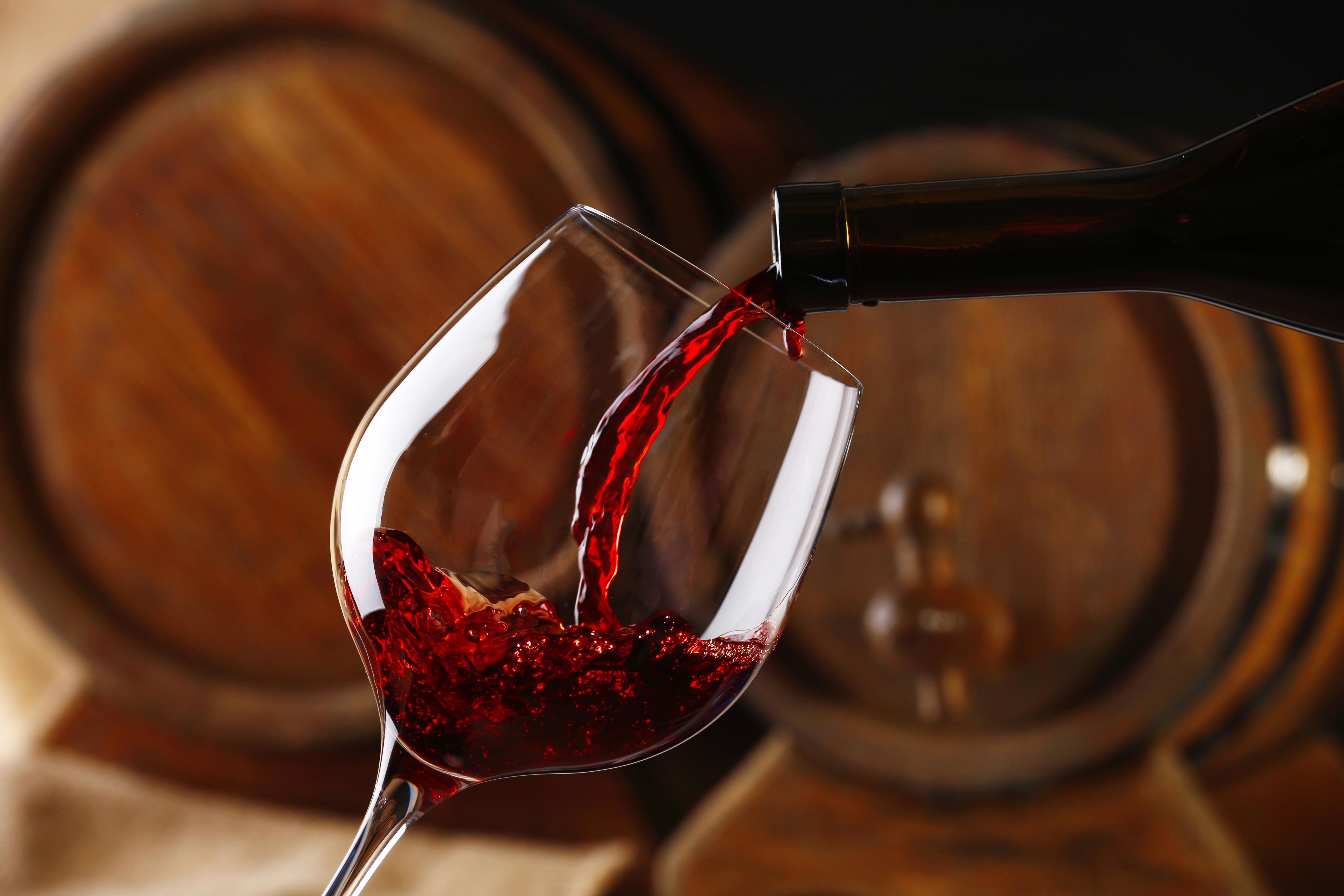 A close-up image of red wine being poured from a bottle into a tilted wine glass. The background features blurred wooden barrels, suggesting a winery or cellar setting. 